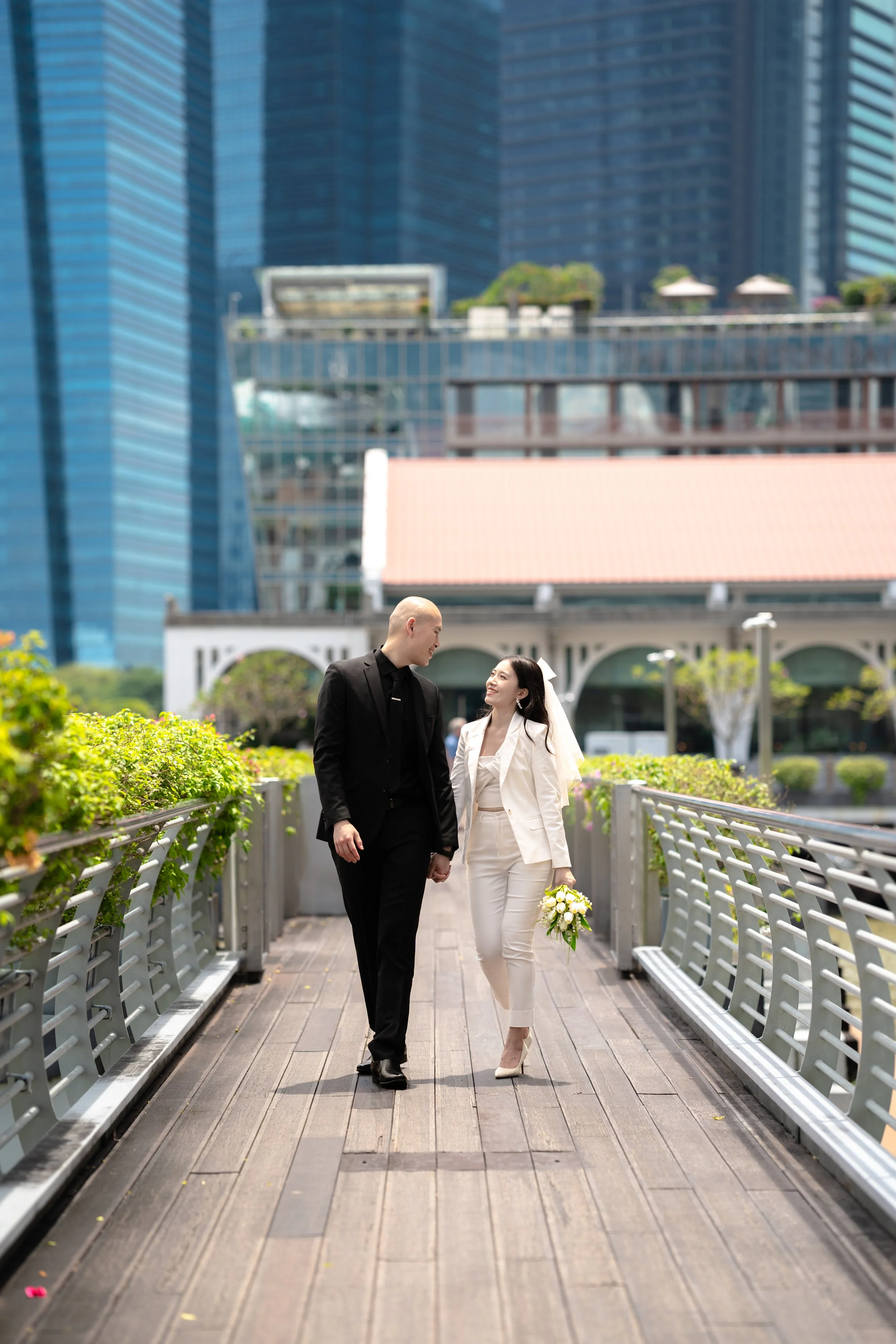 A bride and groom walking hand-in-hand on a wooden bridge in an urban setting, surrounded by modern glass buildings, with the bride holding a bouquet of white flowers and both smiling at each other.