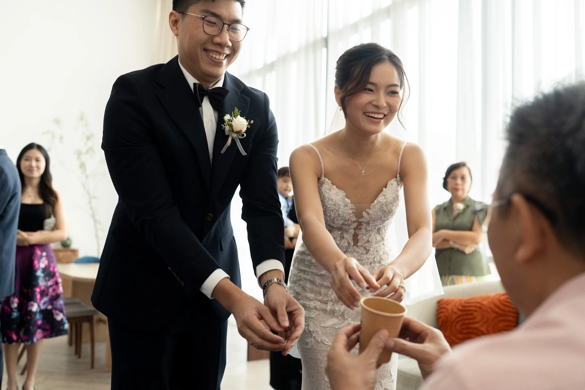 Bride and groom exchanging gifts with an elderly woman during a wedding celebration, with several guests in the background.