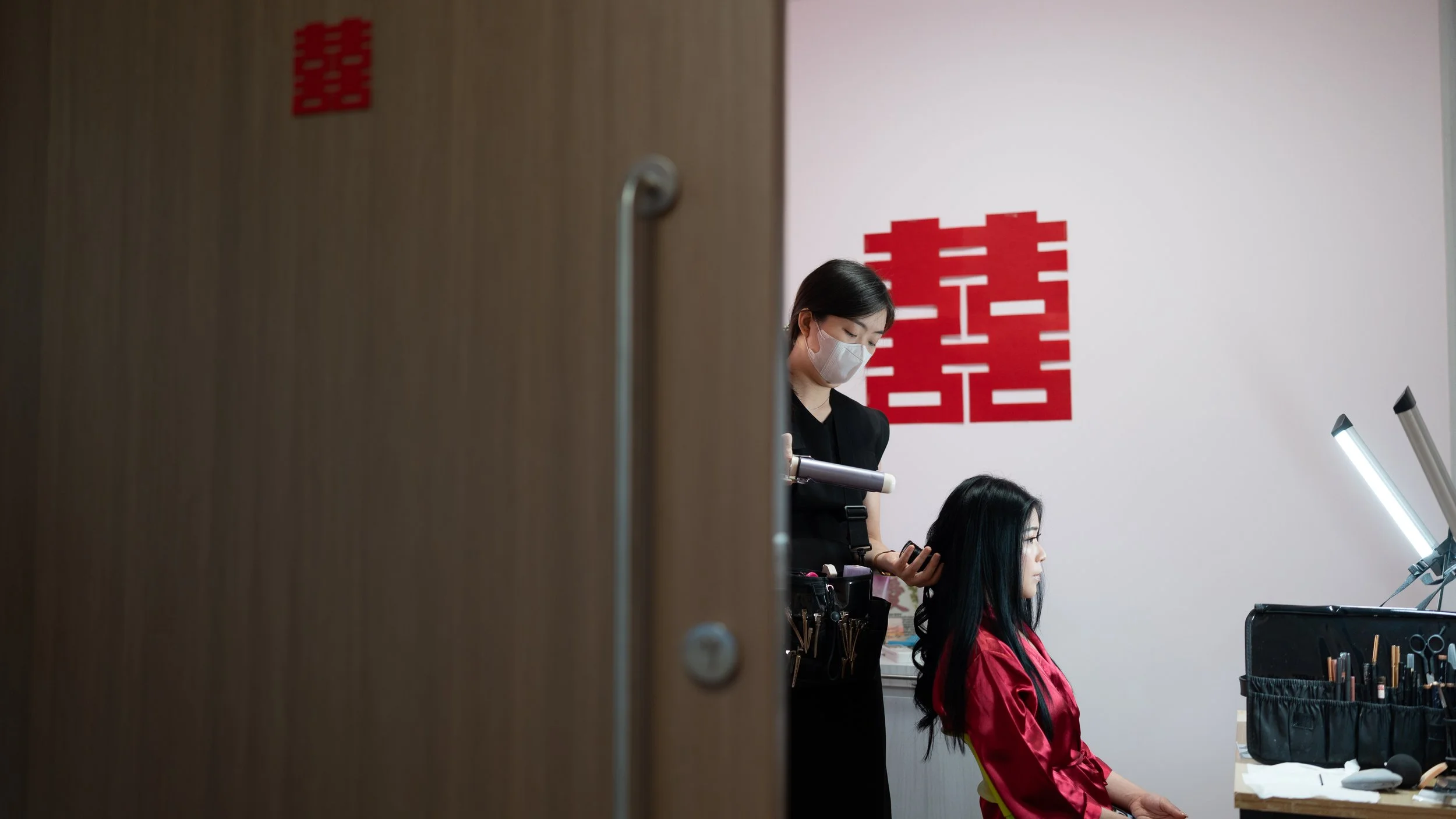 A hairstylist wearing a face mask and black outfit styling a woman's long black hair in a salon. The woman, dressed in a red cape, is seated in front of a mirror with professional hair styling tools on the table.
