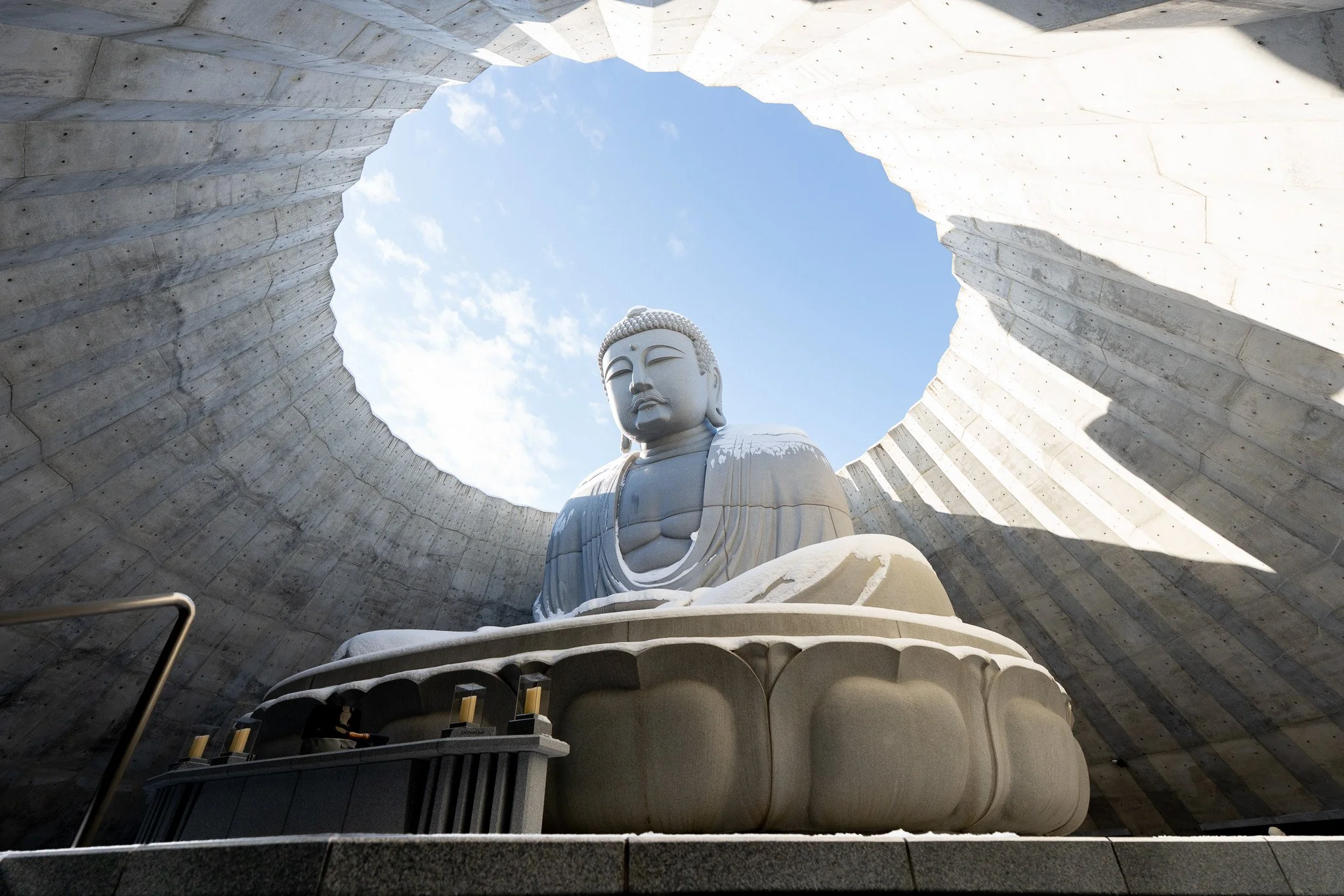 Large white Buddha statue sitting in a hollowed-out concrete structure with a circular opening at the top revealing a blue sky with clouds.