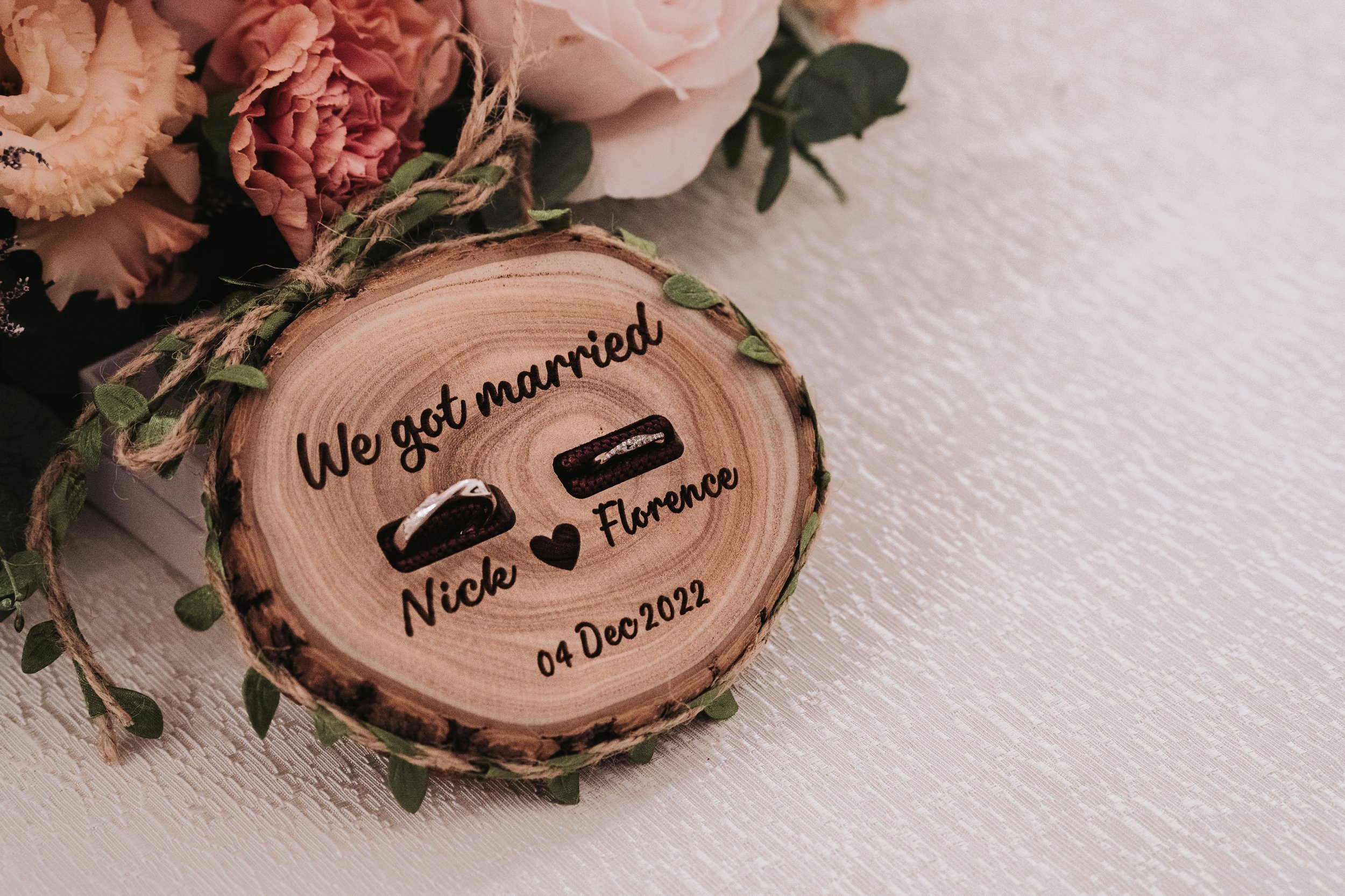 Wooden slice with engraved text celebrating a marriage, surrounded by flowers on a white textured surface.