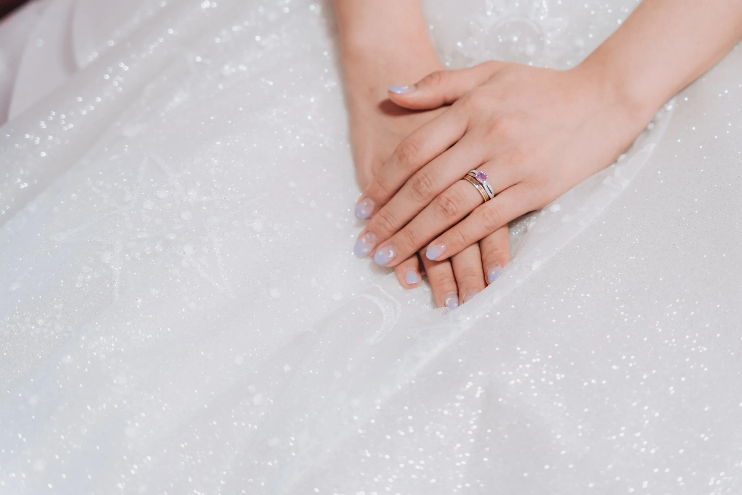 Close-up of a bride's hands with wedding rings, resting on her wedding dress with glitter and sparkles.