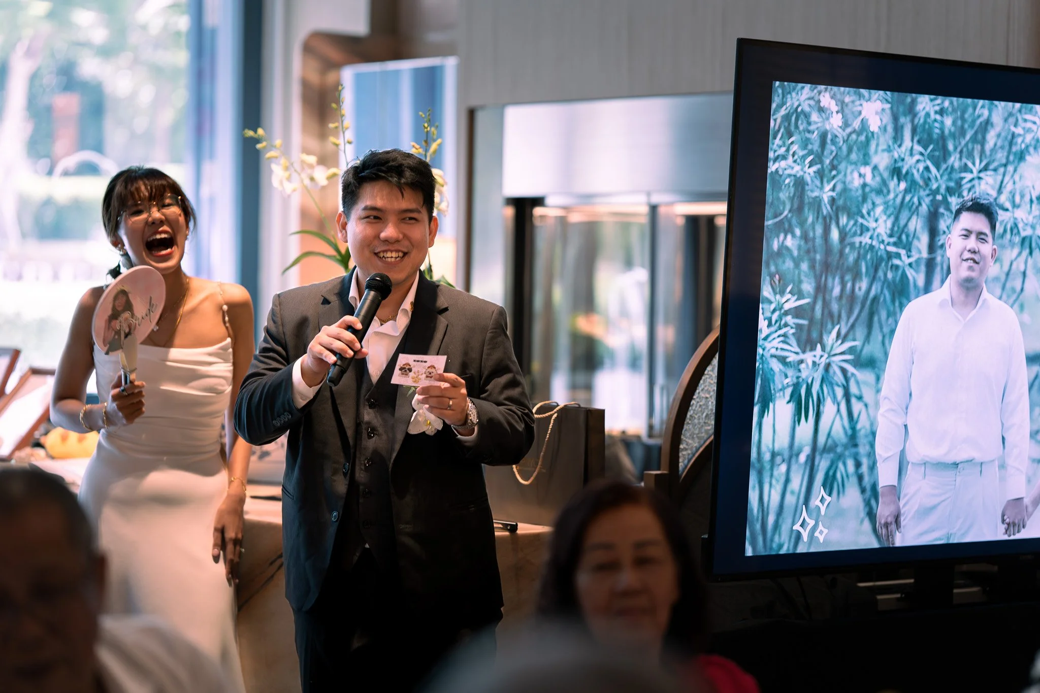 A man in a suit holding a microphone and a photo, smiling, standing next to a woman in a white dress holding a fan, laughing, at an indoor event with a large screen displaying a photo of another man in white shirt and pants.
