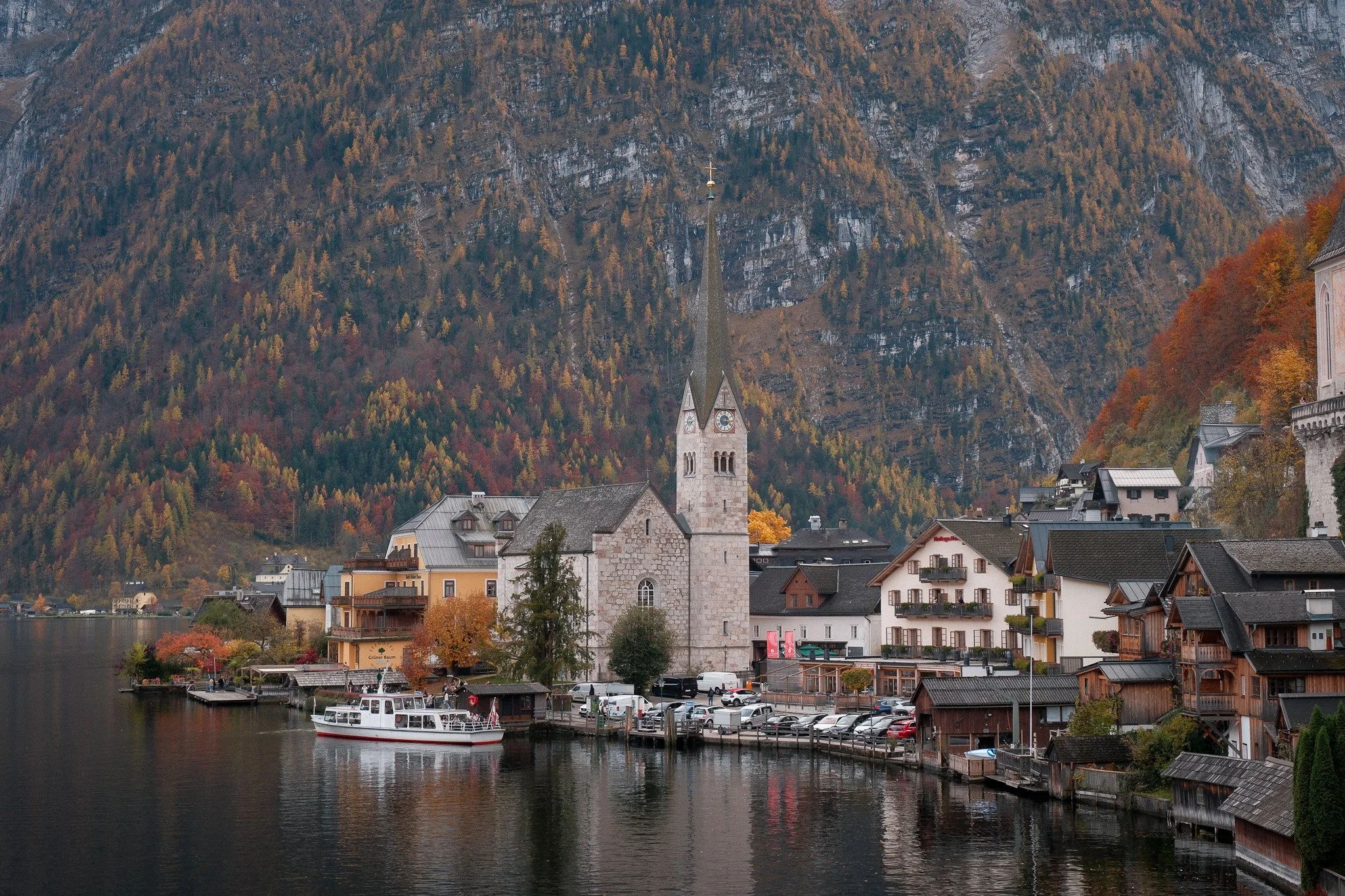 A lakeside village with colorful autumn trees on the hills, a church with a tall spire, parked cars, and a boat on the water.
