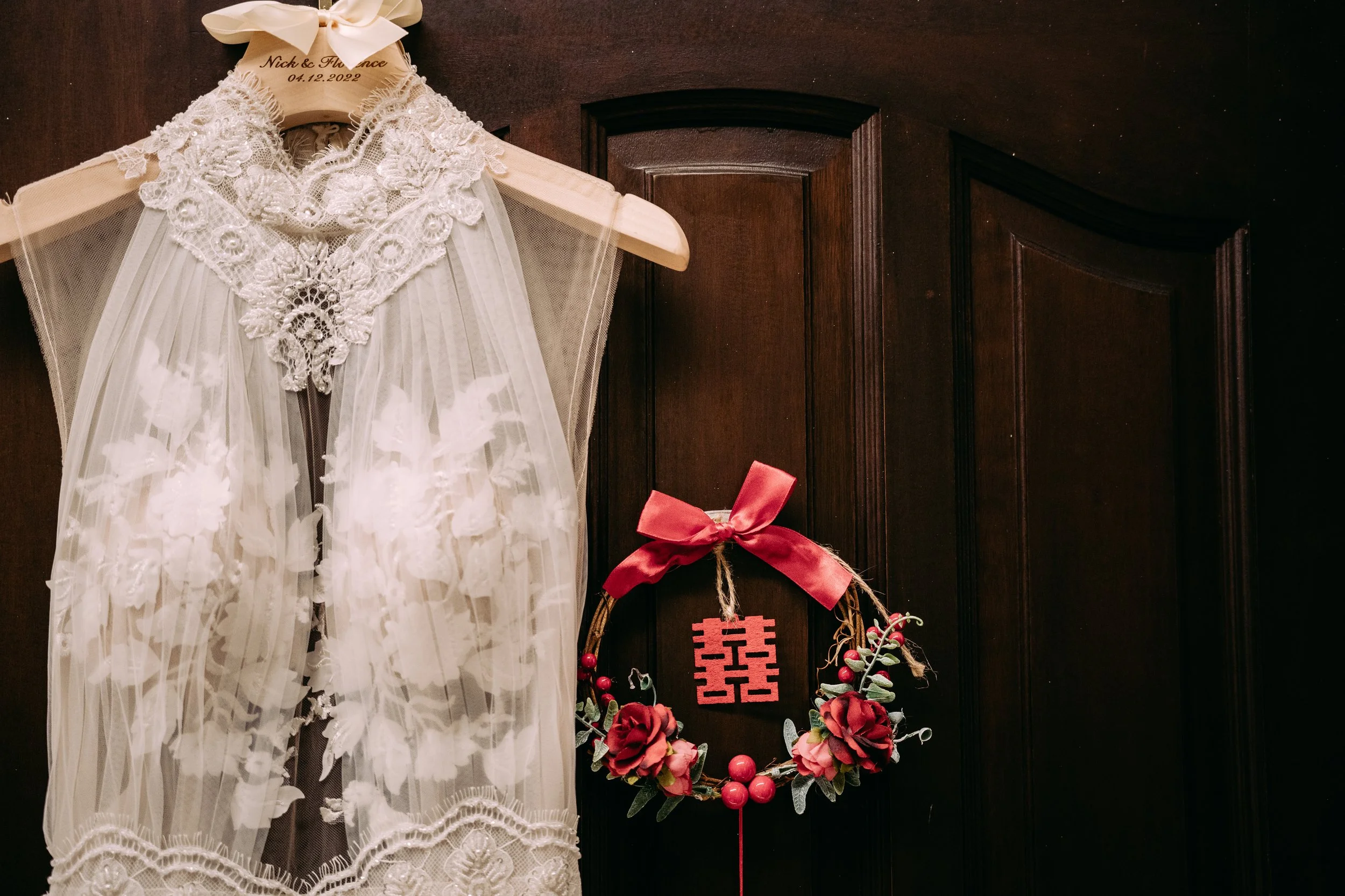 A lace wedding veil with floral embroidery hanging on a wooden door, decorated with a red ribbon bow and a floral wreath with roses and berries, featuring a double happiness Chinese character.