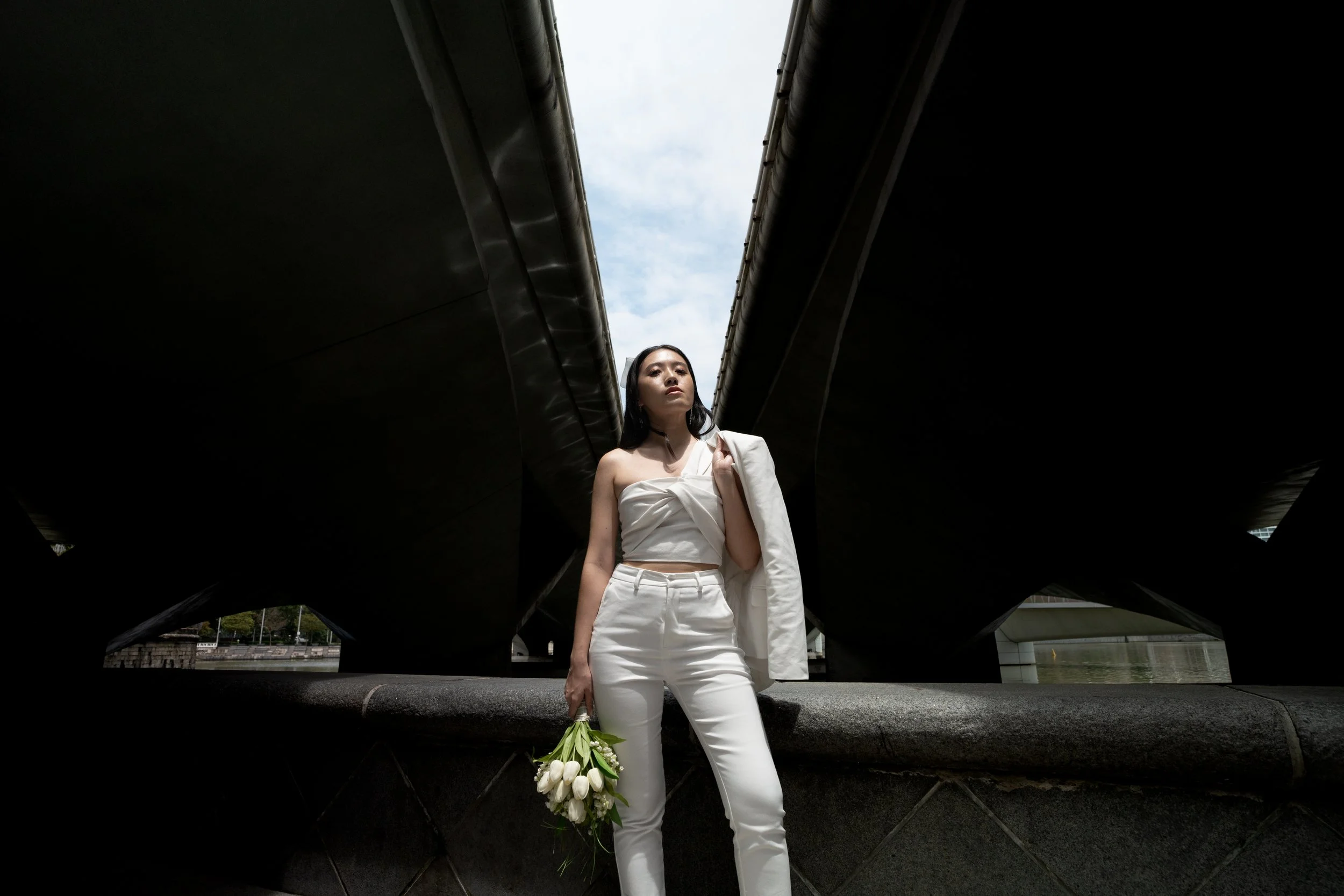 A woman dressed in white holding a bouquet of white tulips, standing outdoors between two large, dark bridge supports with a cloudy sky overhead.