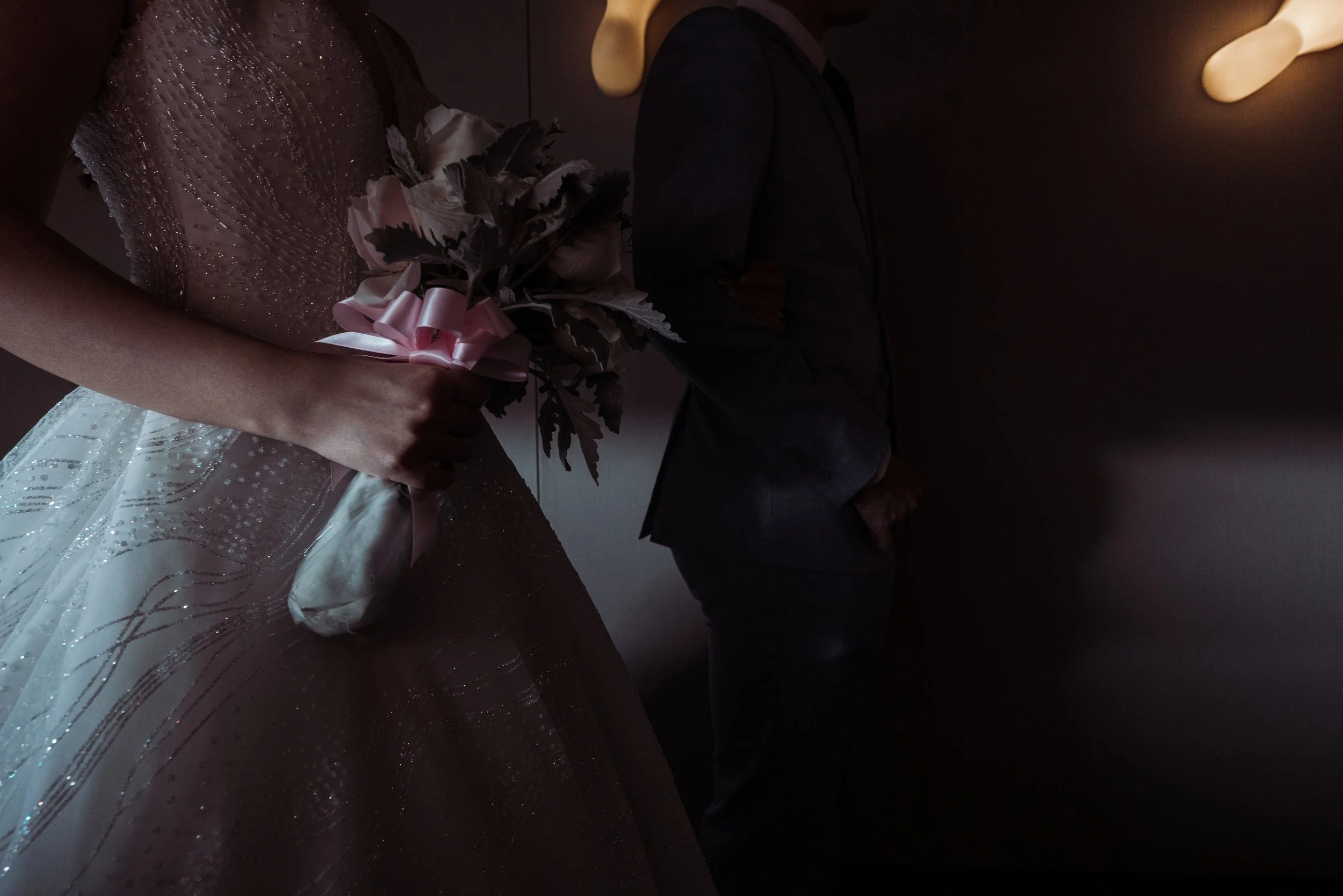 A bride holding a bouquet of flowers in a wedding gown standing next to a person in a dark suit with a cowboy hat, in a dimly lit setting.