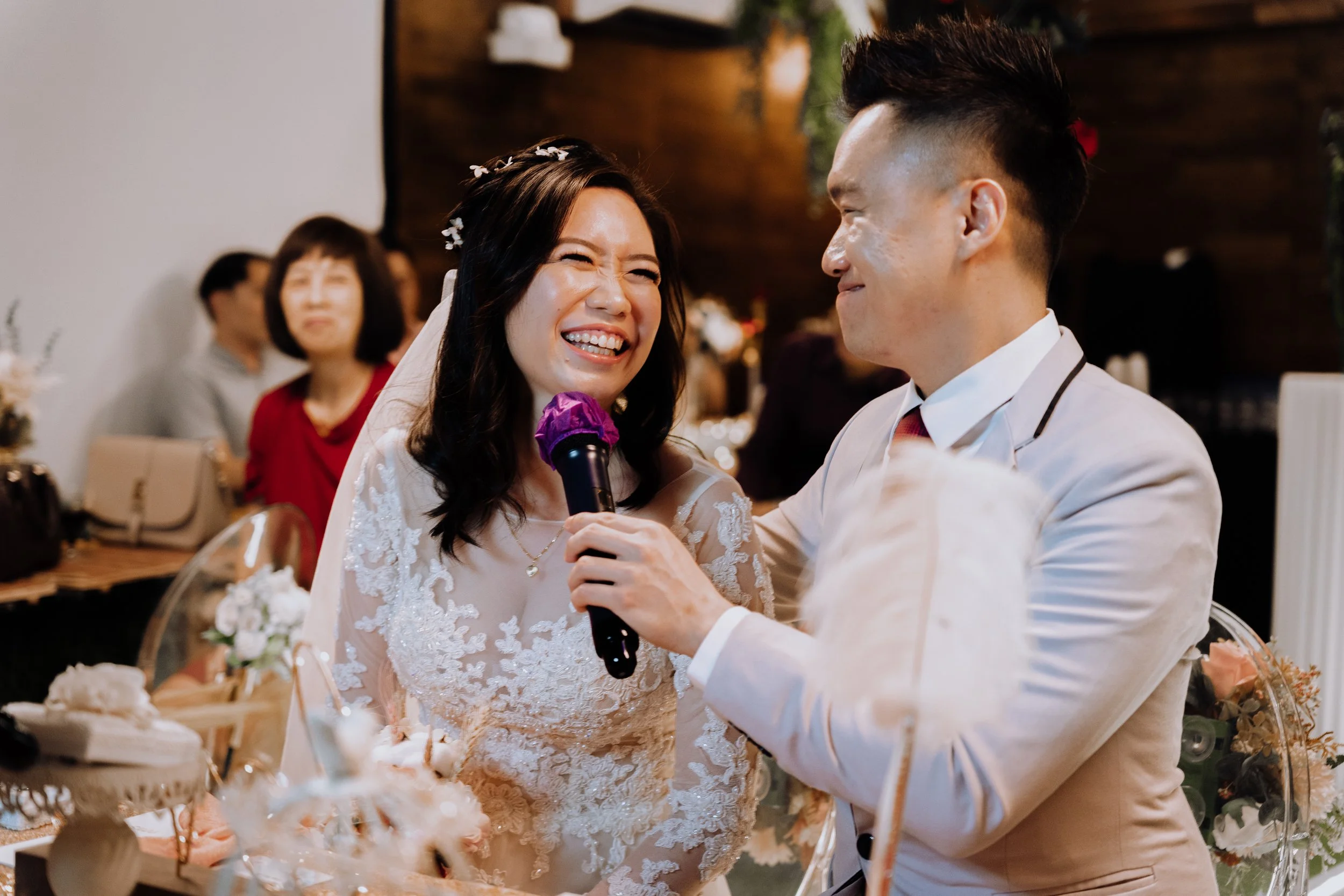A bride and groom laughing and smiling during their wedding ceremony, with a woman in the background watching, in an indoor wedding venue decorated with flowers.