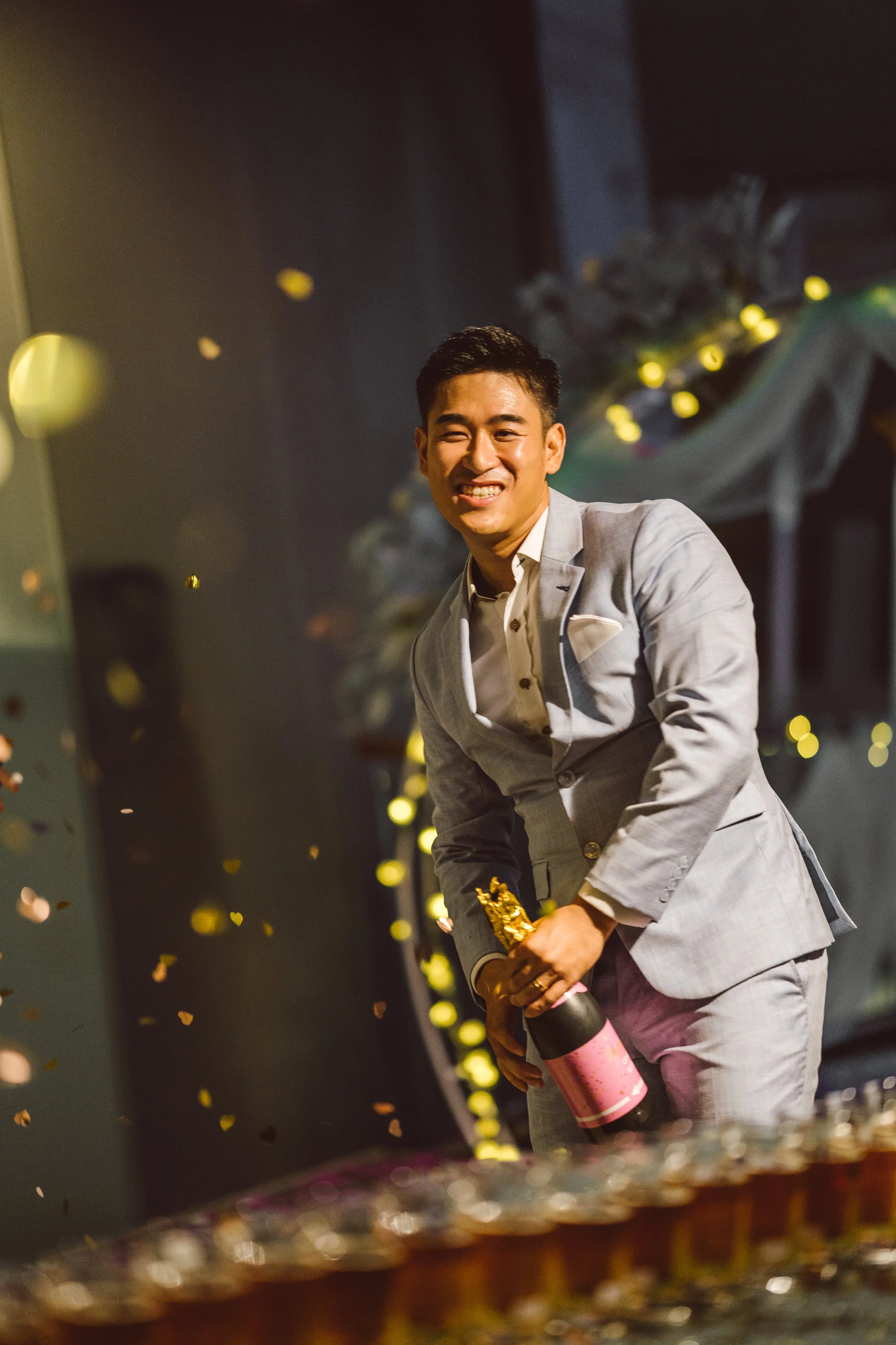 A man in a gray suit holding a bottle of champagne at a celebration event with confetti falling around him.