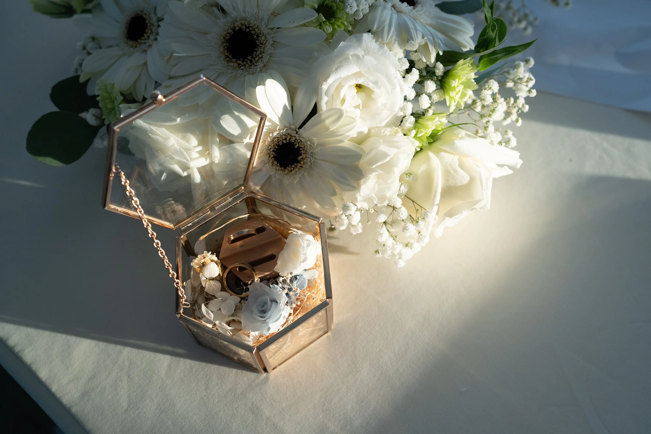A collection of white flowers, including daisies, roses, and baby's breath, next to a glass jewelry box containing rings and a chain, on a cream-colored surface.