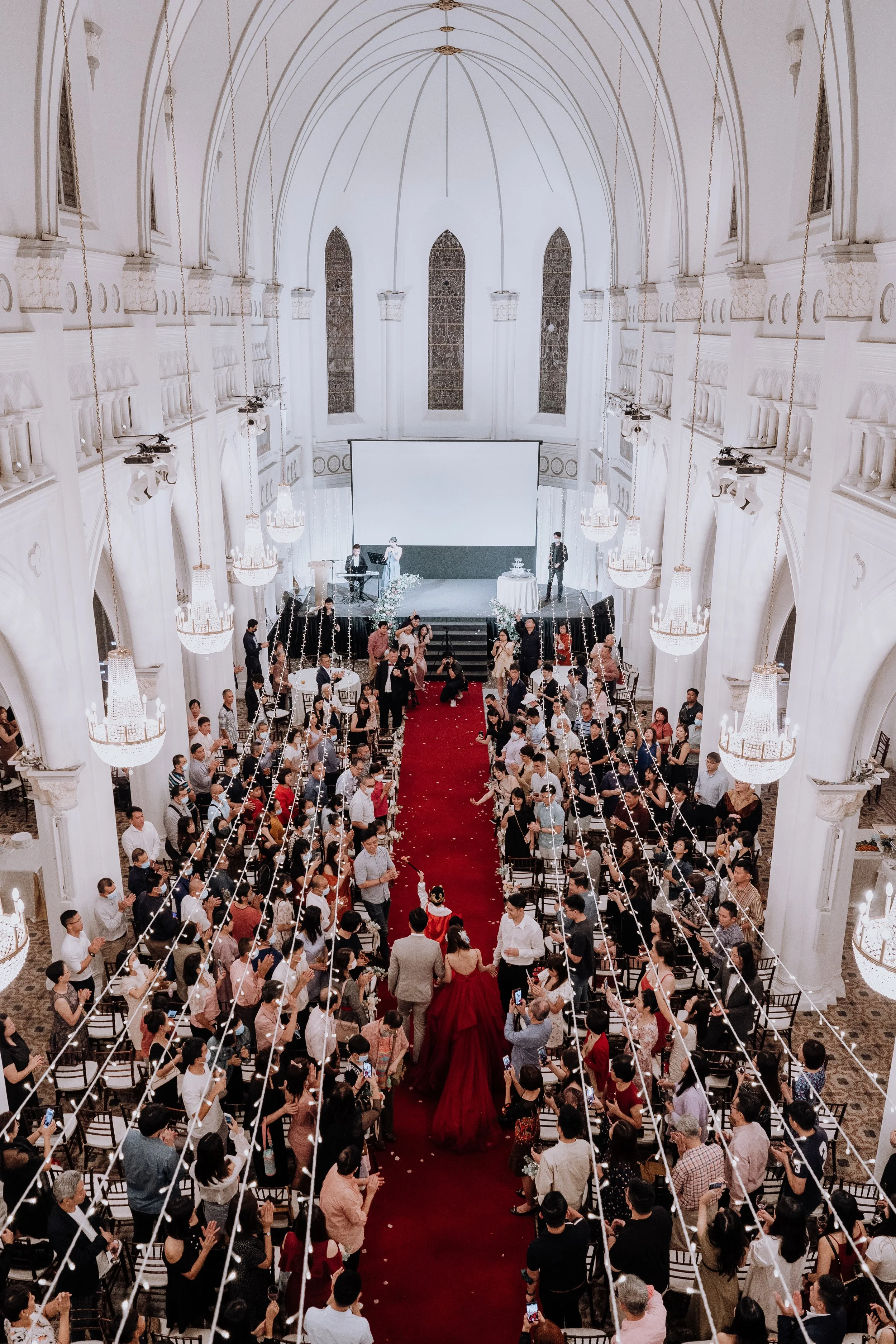 A large wedding or celebration ceremony inside a grand, white cathedral-style hall with high arched ceilings, stained glass windows, chandeliers, and a red carpet aisle. Guests are gathered around, some taking photos, as the bride and groom walk down