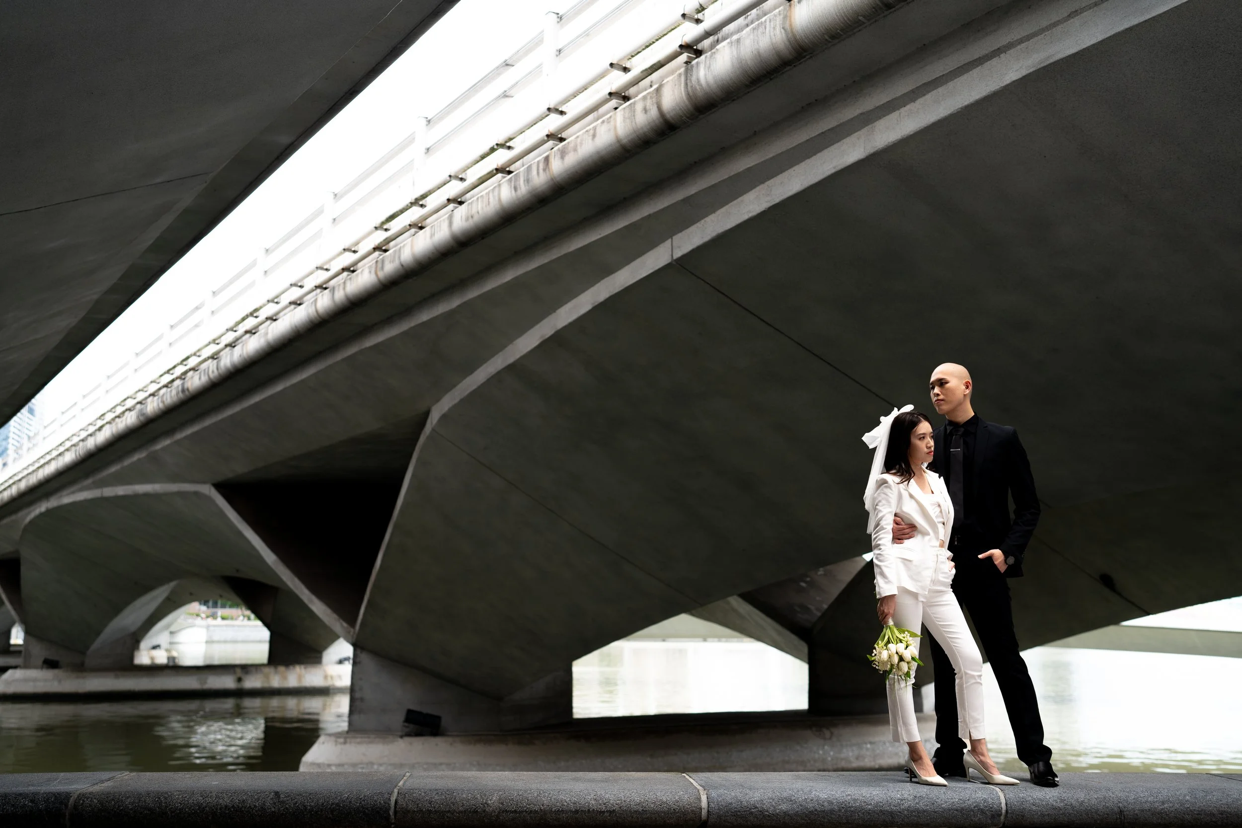 A couple in wedding attire stands under a modern bridge near a body of water, holding a bouquet of flowers.
