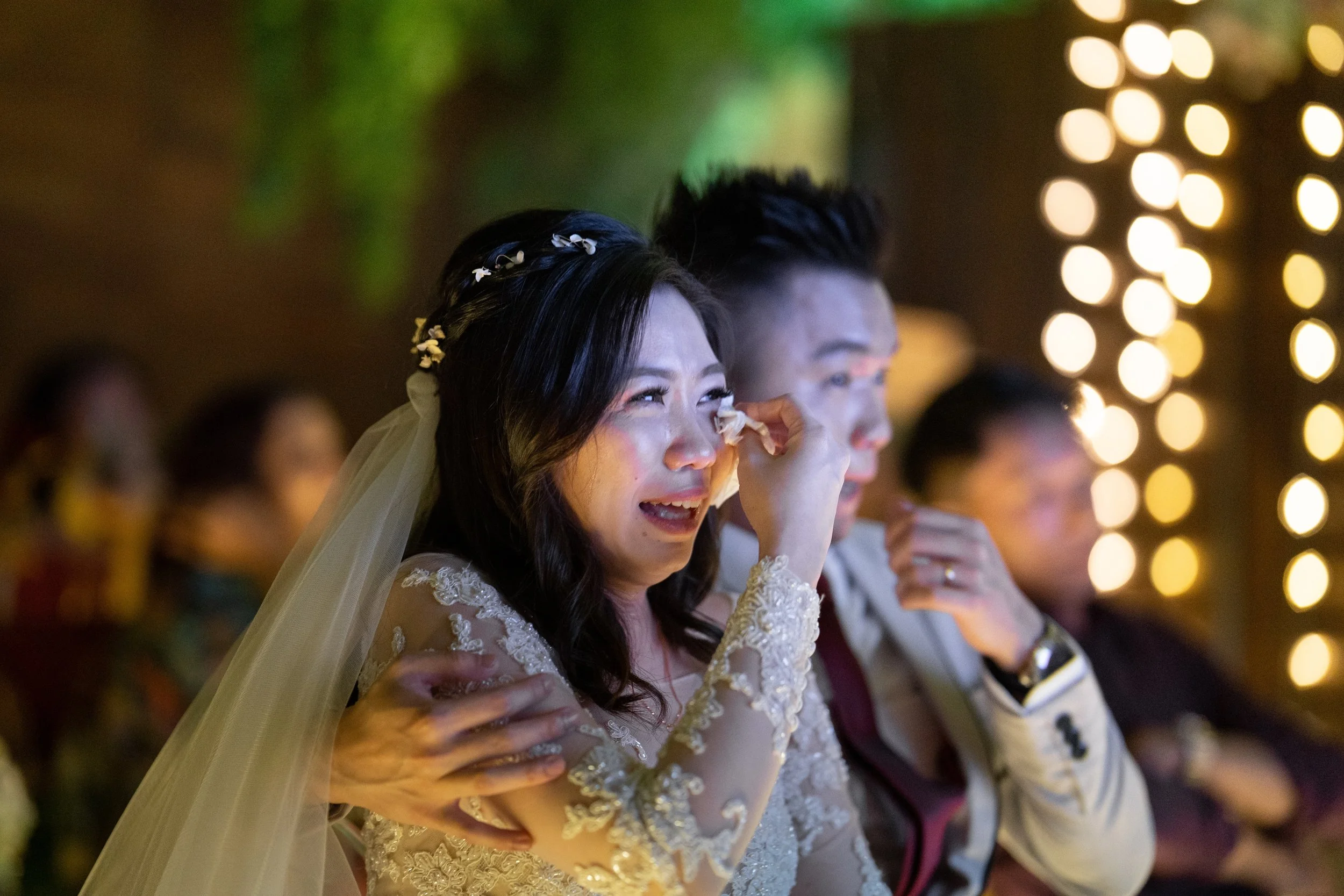 A bride crying and wiping tears with a tissue during her wedding reception, seated beside her groom and other guests, with decorative lights in the background.
