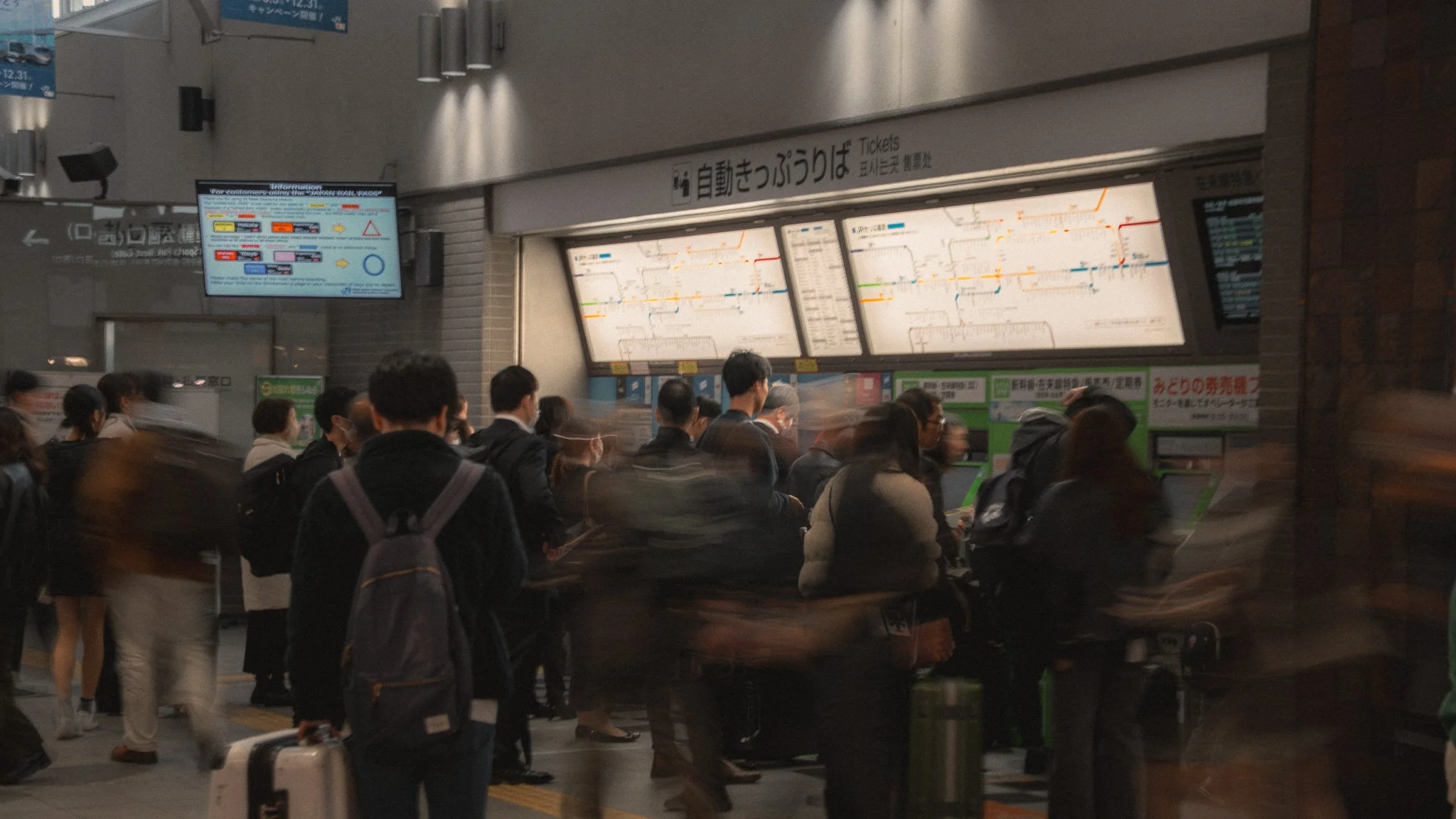 People waiting in line at a ticket counter in a train station, with digital maps and signs overhead.