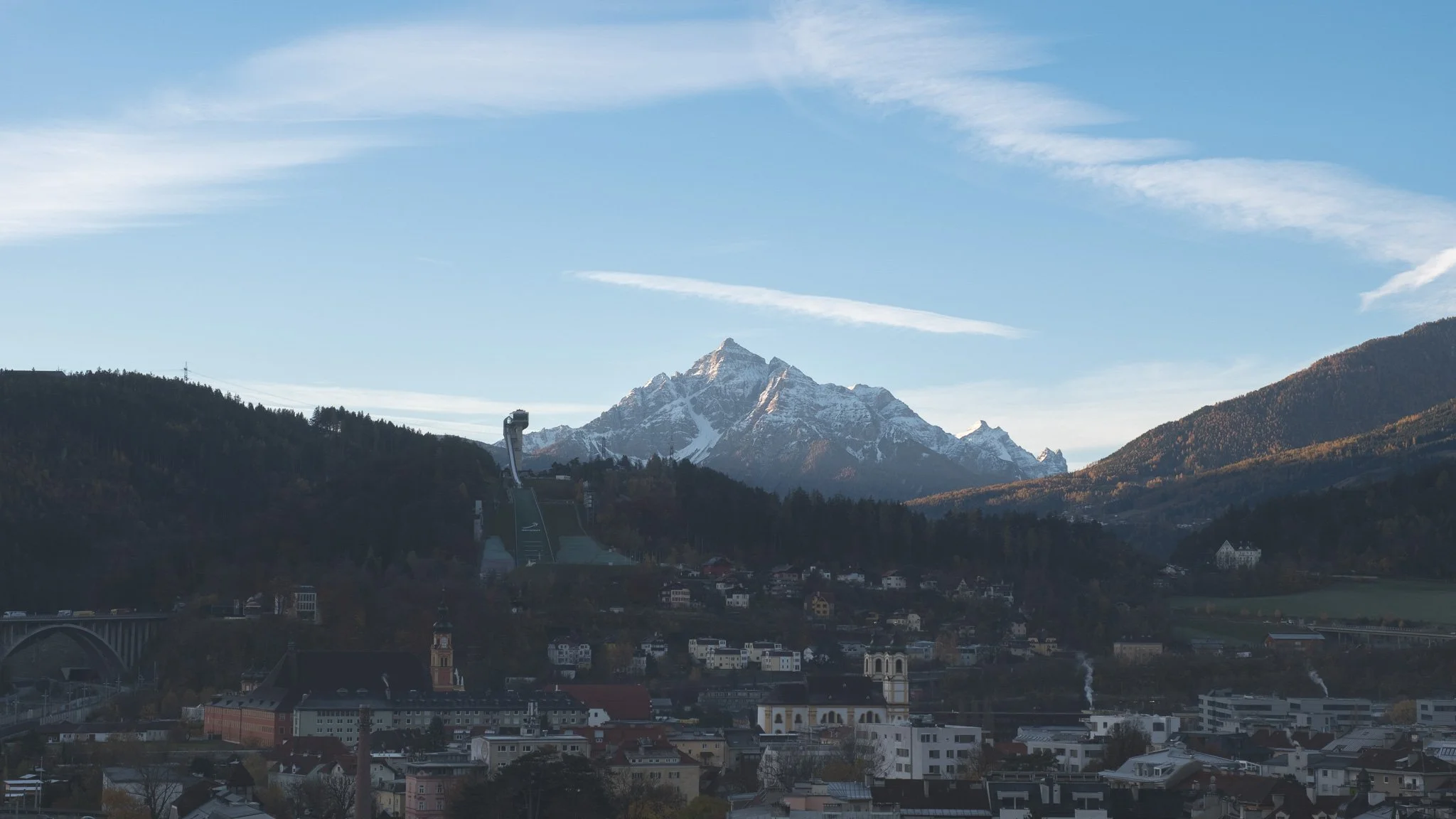 A scenic view of a mountain range with snow-capped peaks, a city with buildings and houses below, and ski jump ramps on the hillside, under a blue sky with some clouds.