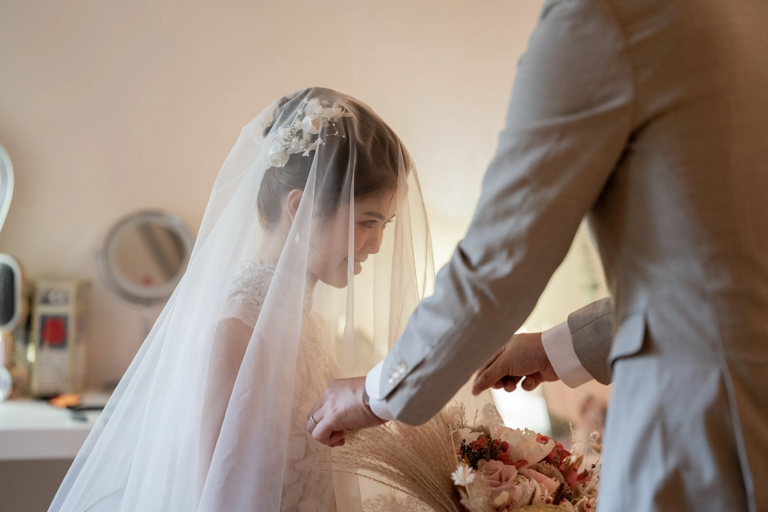 A bride in a wedding gown and veil with floral embellishments, holding a bouquet of pink and white flowers, as a groom in a gray suit interacts with her during a wedding ceremony indoors.