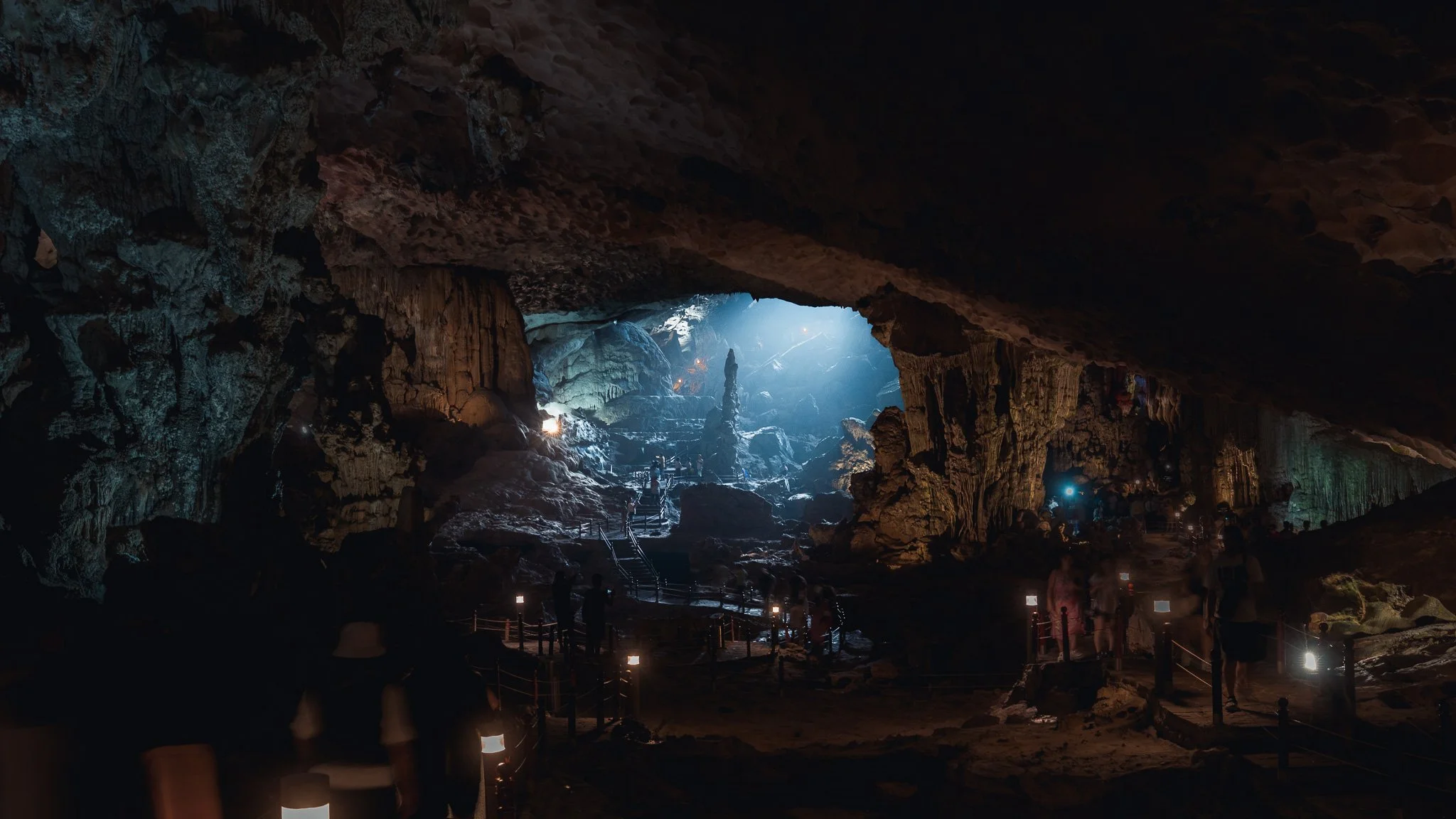 People exploring the dark, illuminated interior of a large cave with stalactites and stalagmites, pathways with lights and railings.