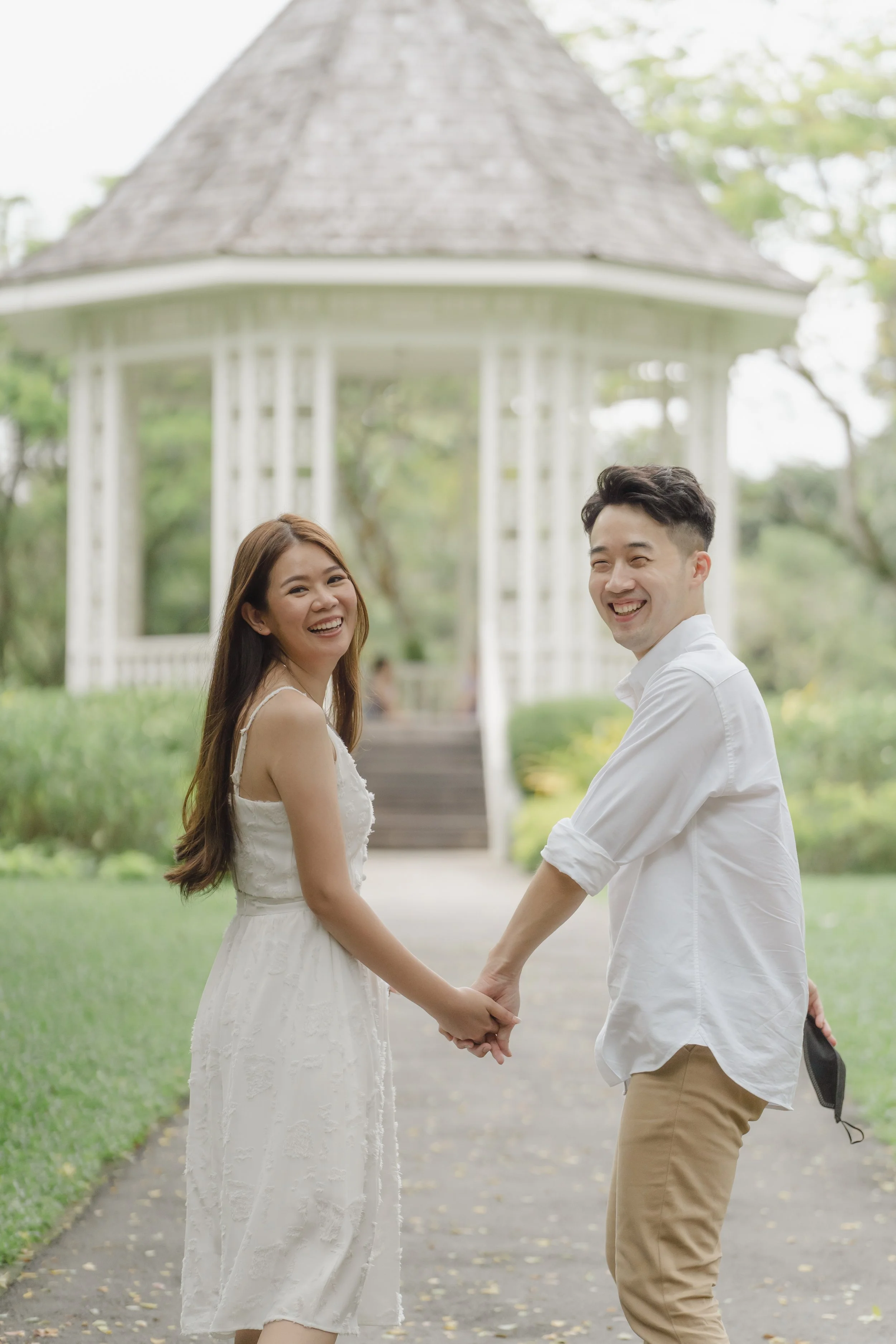 A couple holding hands and smiling outdoors in front of a gazebo, with trees and greenery in the background.