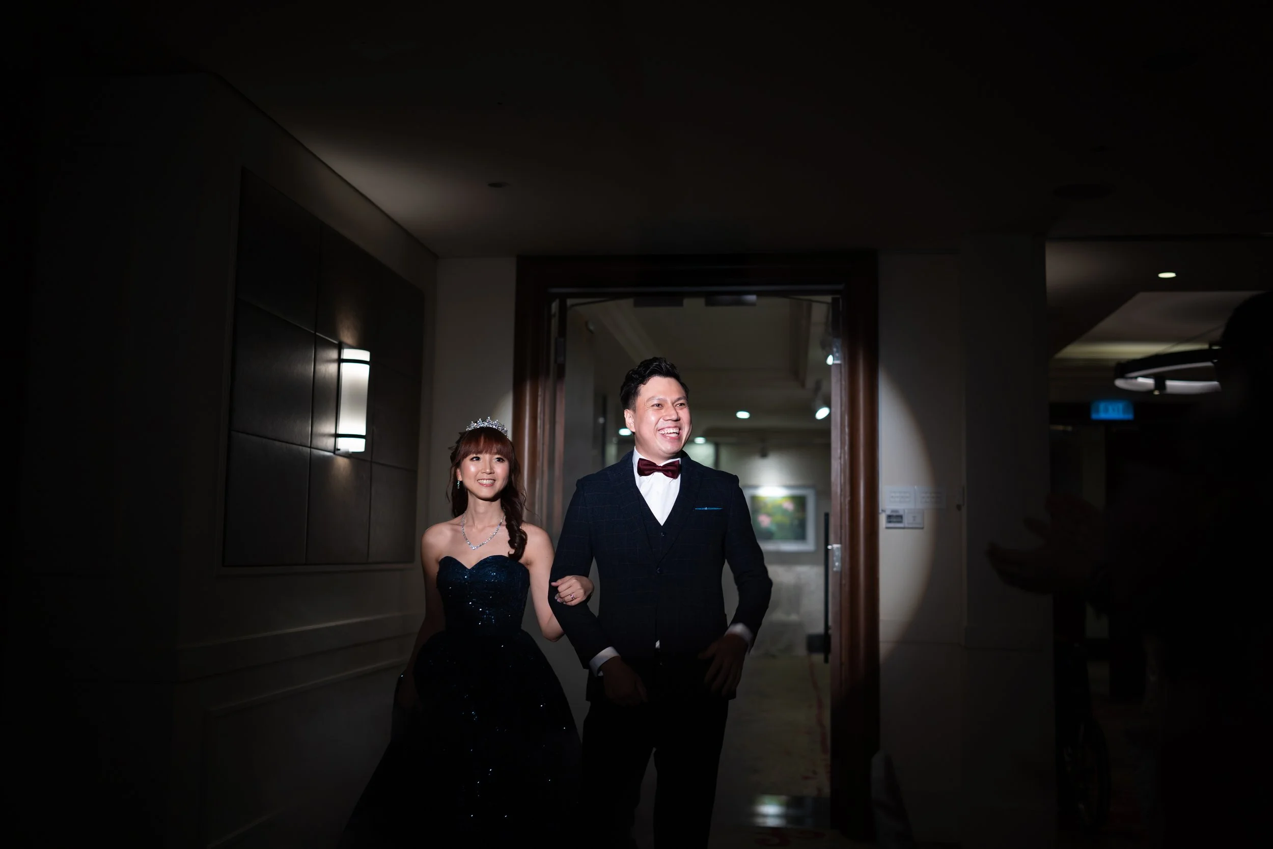 A happy wedding couple walking arm in arm through a dimly lit hallway. The bride is wearing a dark strapless gown with a tiara and necklace, and the groom is in a dark suit with a bow tie. They are smiling and looking forward.