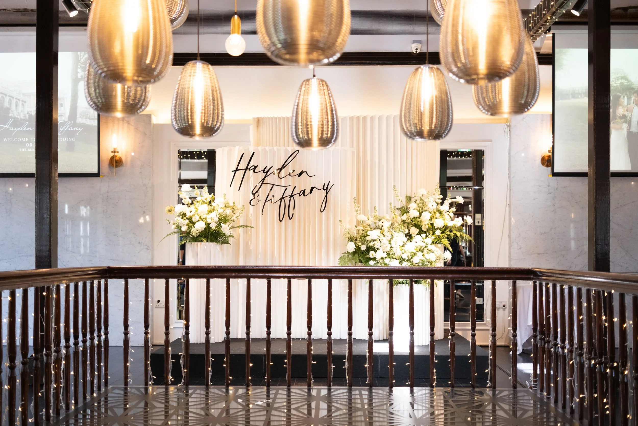 Elegant wedding reception area with white floral arrangements, gold hanging lights, and a backdrop with the names Hayden and Tiffany, featuring a black engraved sign.