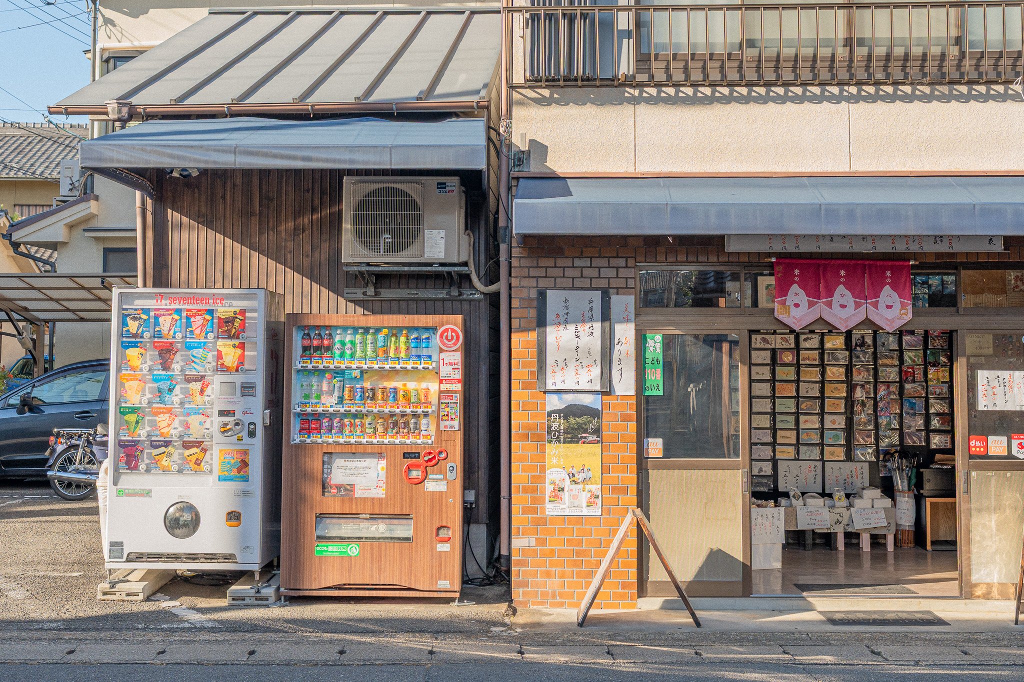 Street view of a small shop with vending machines outside, one for snacks and drinks, and a brick building with a display of mailboxes and a pink hanging banner inside.
