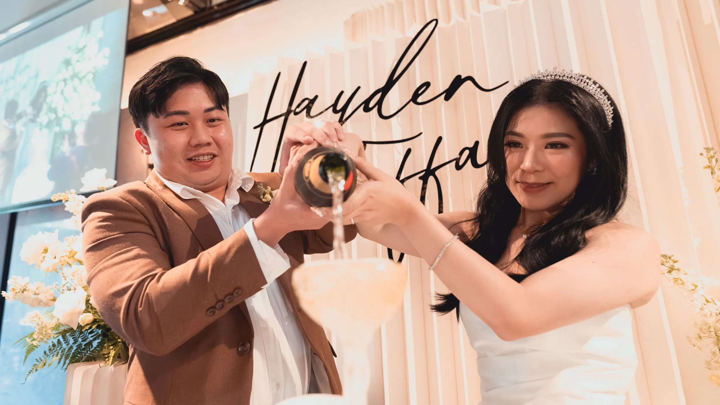 A couple celebrating at their wedding, with the groom pouring champagne into the bride's glass, smiling and looking at each other, standing in front of a decorated backdrop with their names.