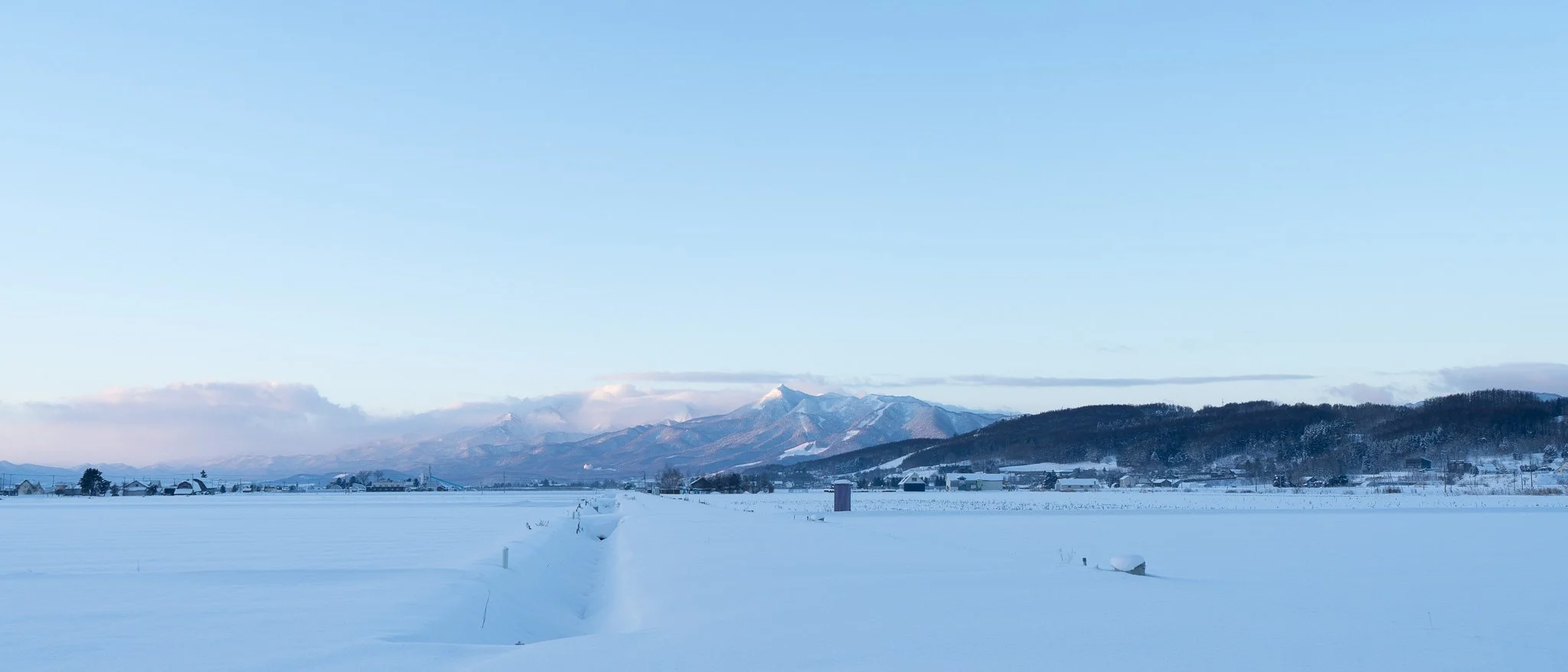 Snow-covered field with a small trail, distant houses, and mountains in the background under a clear blue sky.