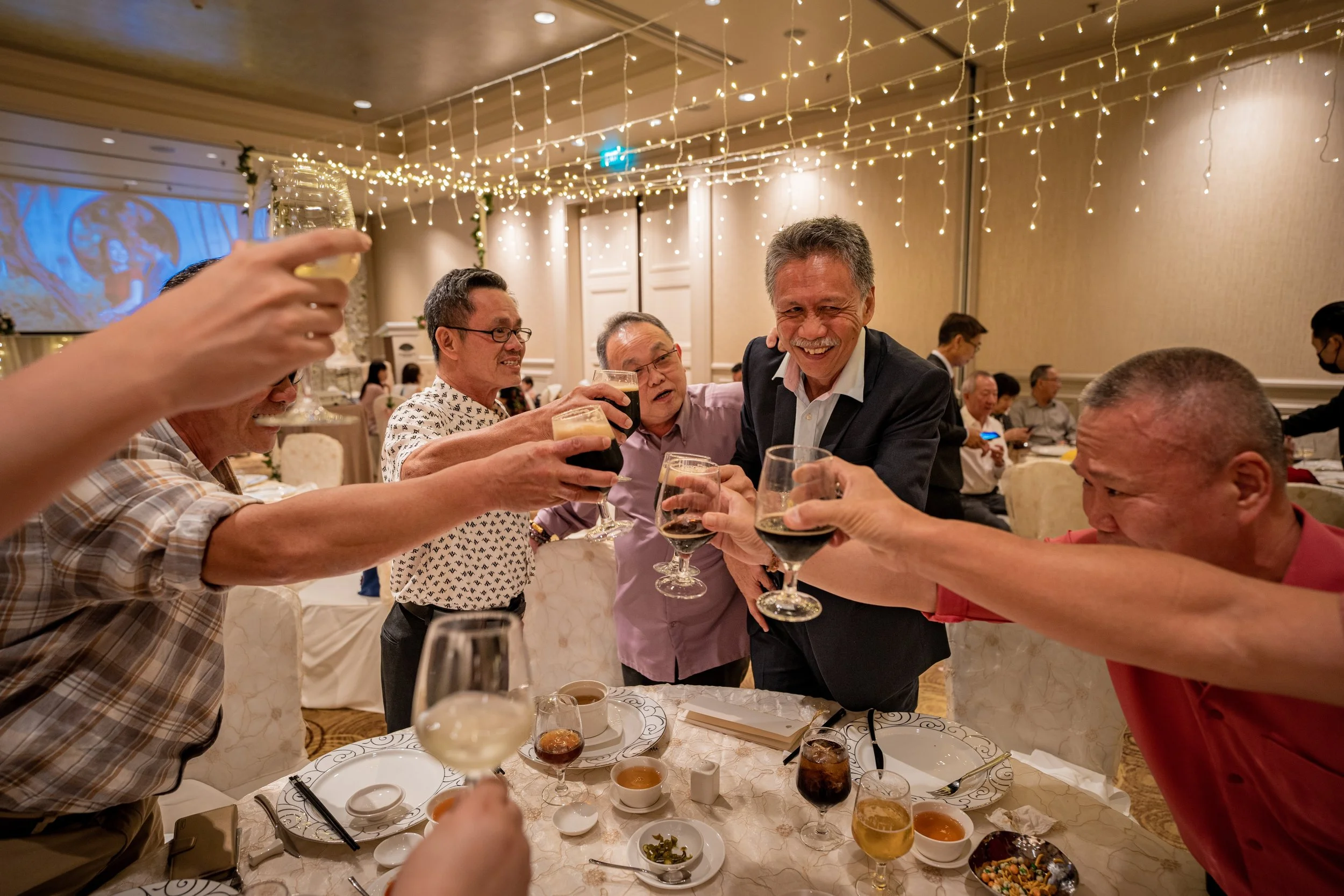 A group of people celebrating at a formal event, raising glasses in a toast, with smiles and laughter, decorated with string lights and elegant table settings.