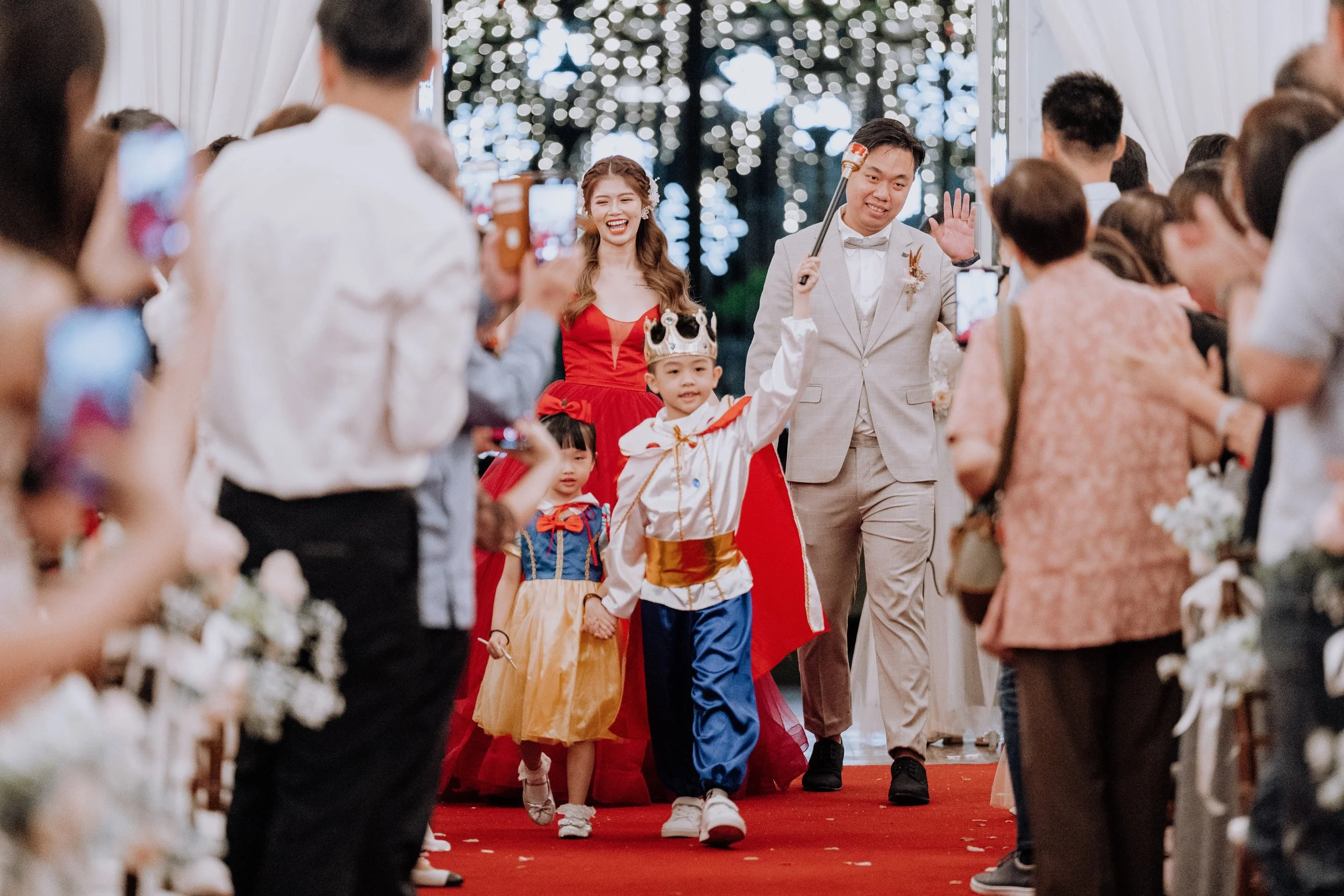 A wedding scene with a bride and groom walking down the aisle, children dressed as royalty, and guests taking photos.