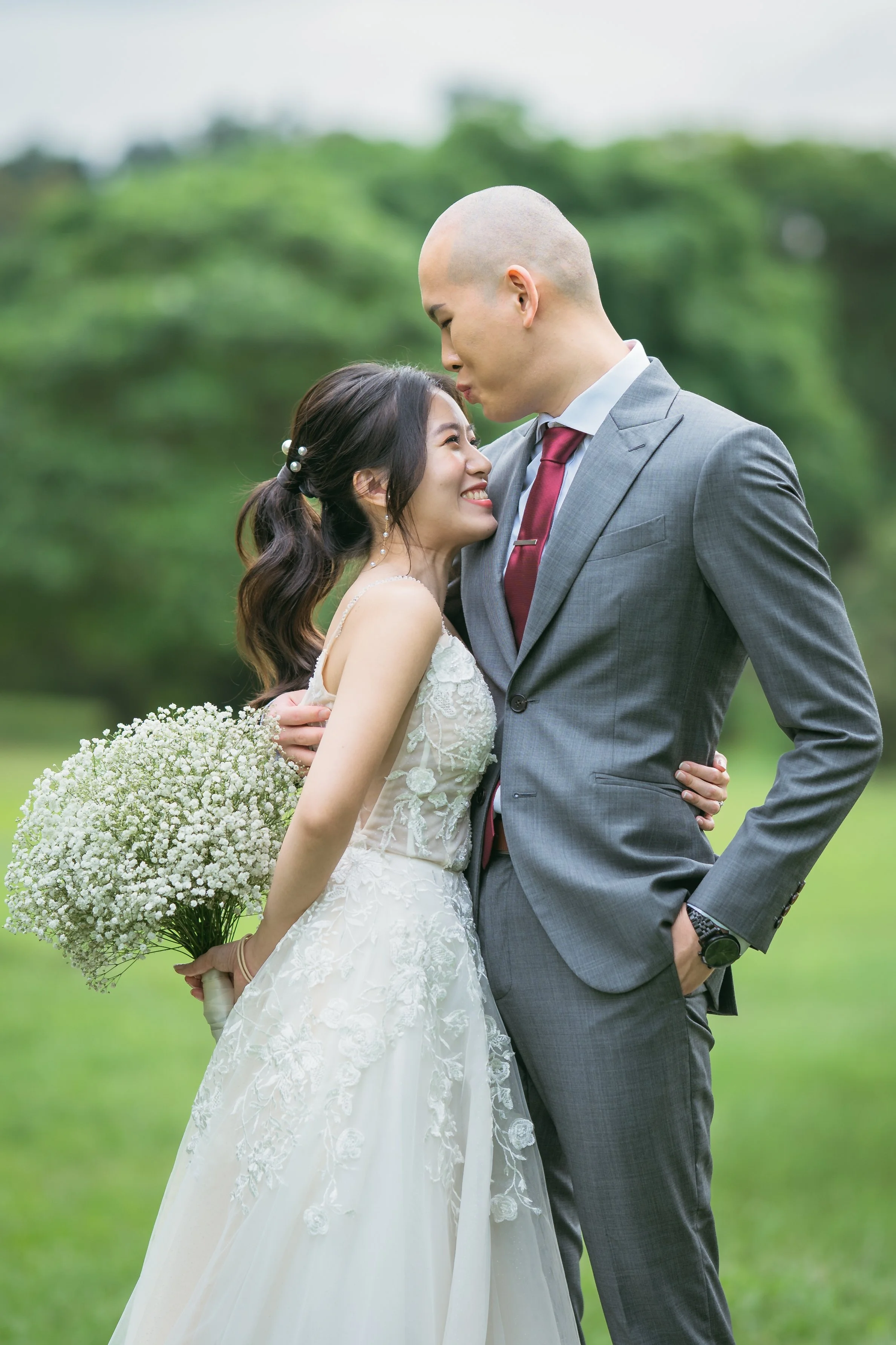 A bride and groom share a loving moment outdoors, with the groom leaning down to touch foreheads, and the bride smiling while holding a bouquet of white baby's breath flowers.