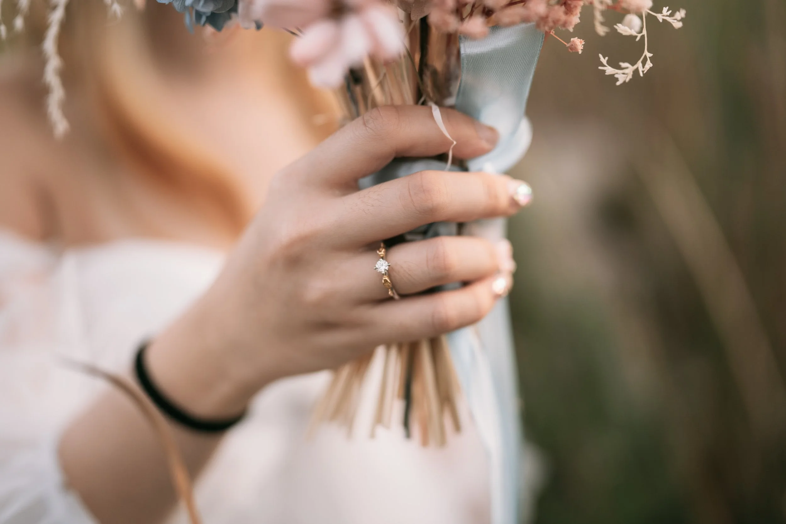 A woman holding a bouquet of flowers with a diamond ring on her finger, visible nails painted with a glittery polish, and a black hair tie on her wrist.
