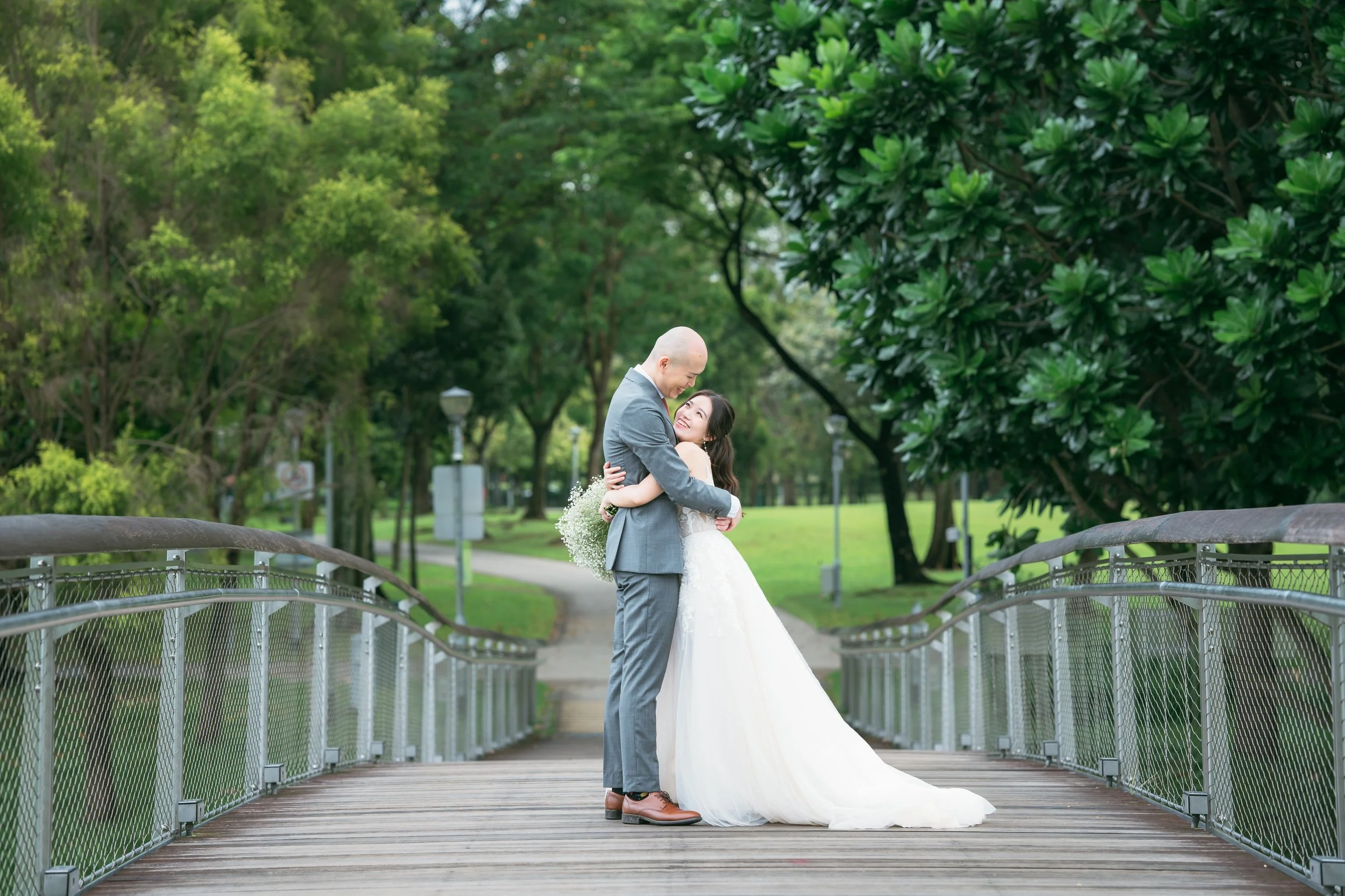 A newlywed couple embracing on a wooden bridge in a park, surrounded by lush green trees.