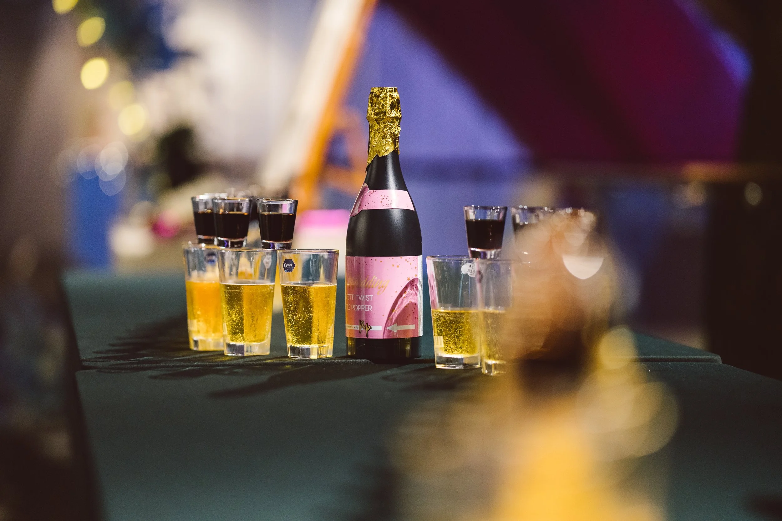A bottle of champagne with a pink label and gold foil on top, surrounded by small glasses of yellow and dark-colored drinks, set on a dark table with a blurred colorful background.
