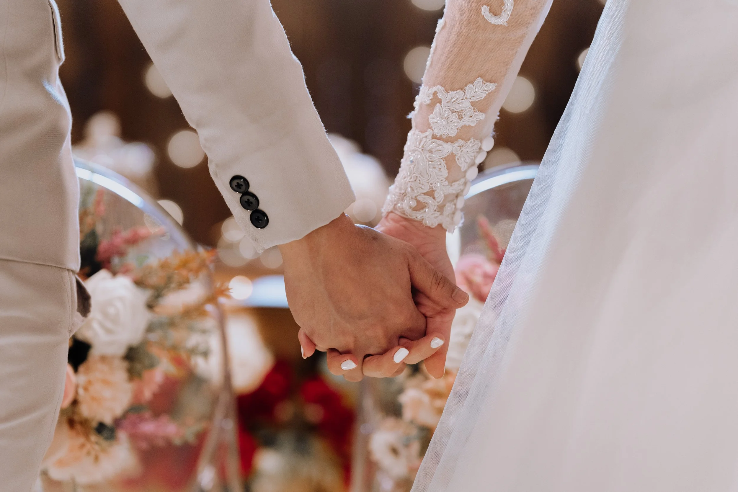 Close-up of a bride and groom holding hands during a wedding ceremony, with a background of flower arrangements and blurred lights.