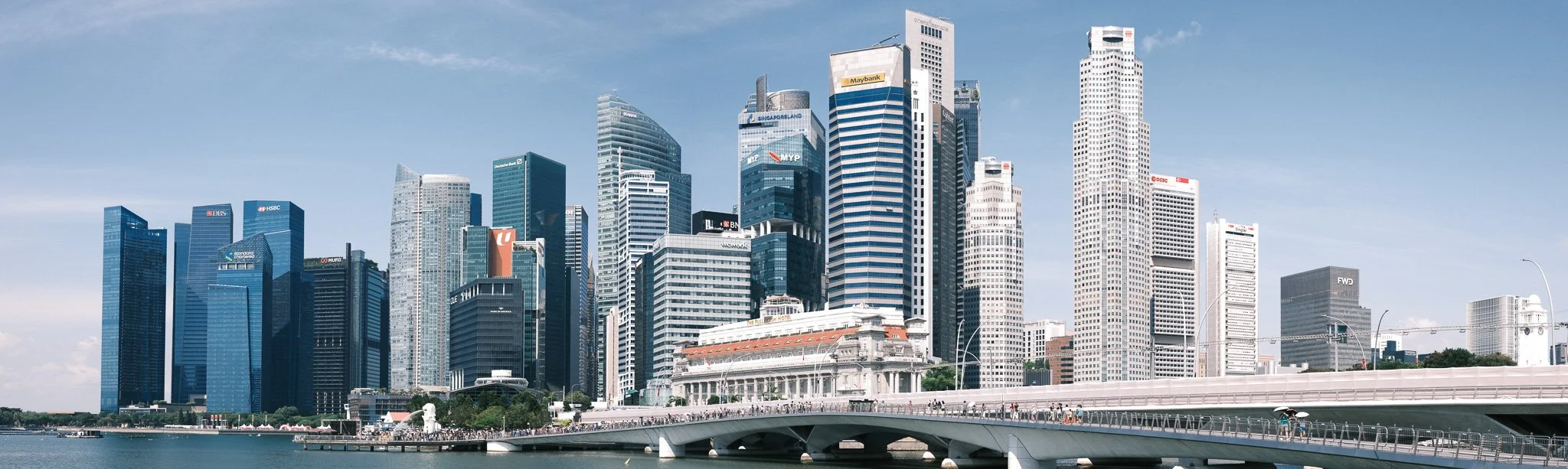 A panoramic view of a modern city skyline with numerous tall skyscrapers, some with corporate logos like HSBC, Maybank, and DBS, seen from across a body of water with a bridge in the foreground.