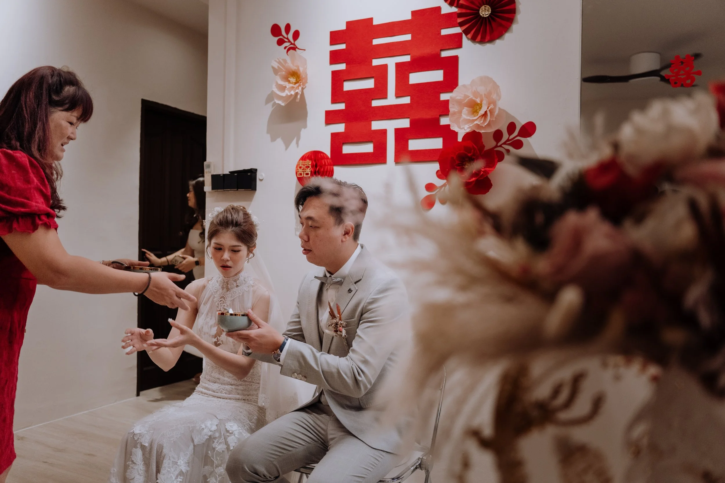 A traditional Chinese wedding tea ceremony, featuring a bride and groom seated and a woman serving tea, with festive red and white decorations on the wall.