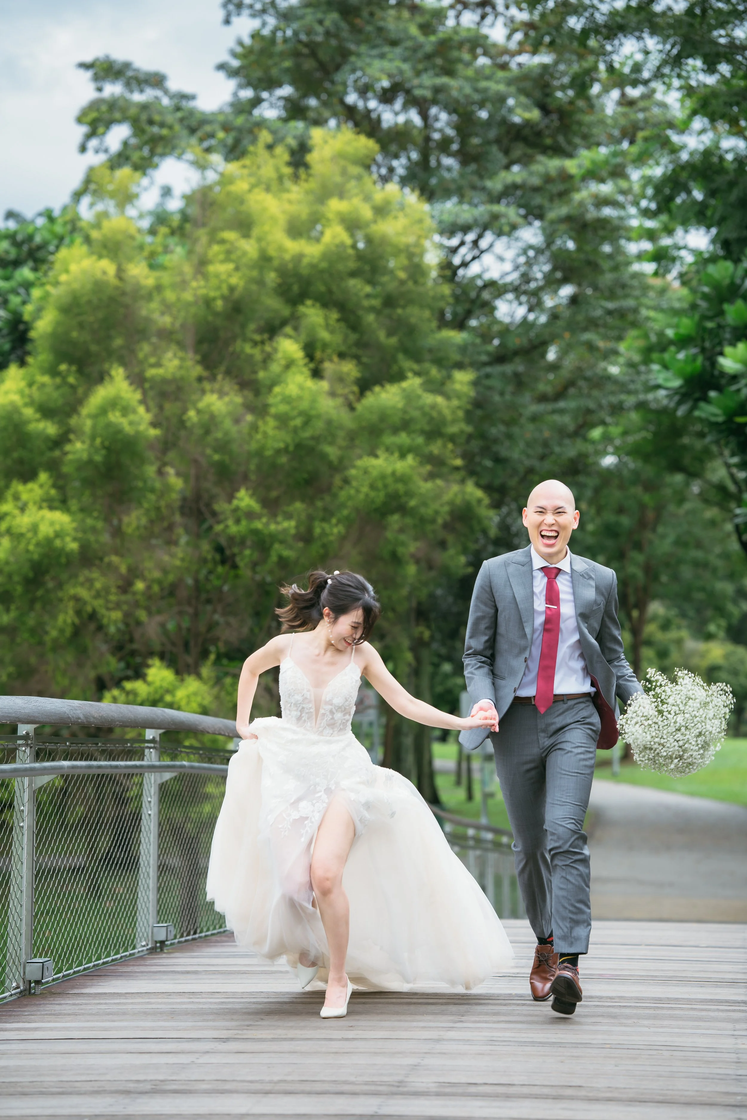 A joyful couple dressed in wedding attire running on a wooden bridge in a park, holding hands and smiling, with lush green trees in the background.
