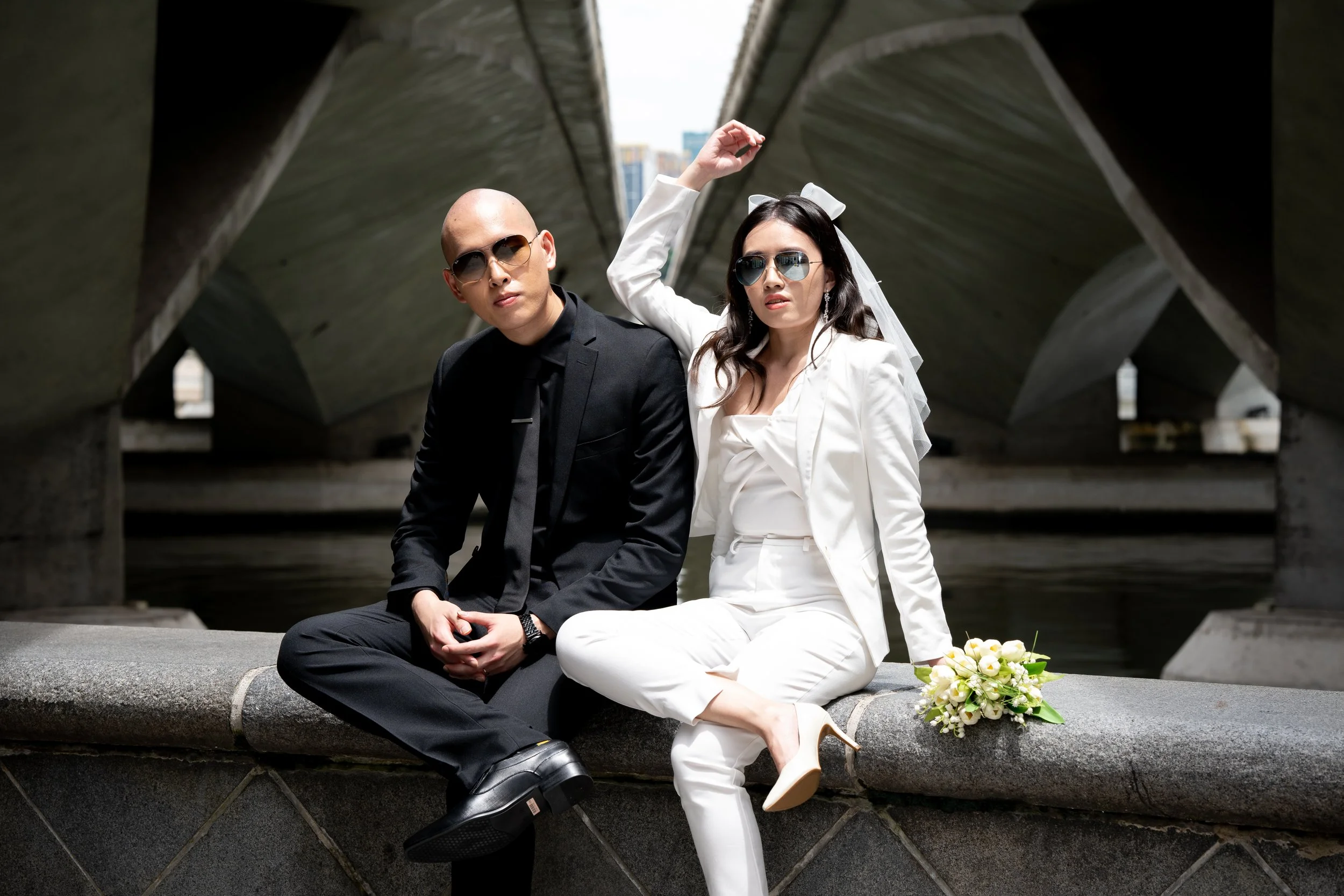 A couple dressed in wedding attire, sitting on a concrete ledge under a bridge, with the woman holding a bouquet of white flowers and both wearing sunglasses.