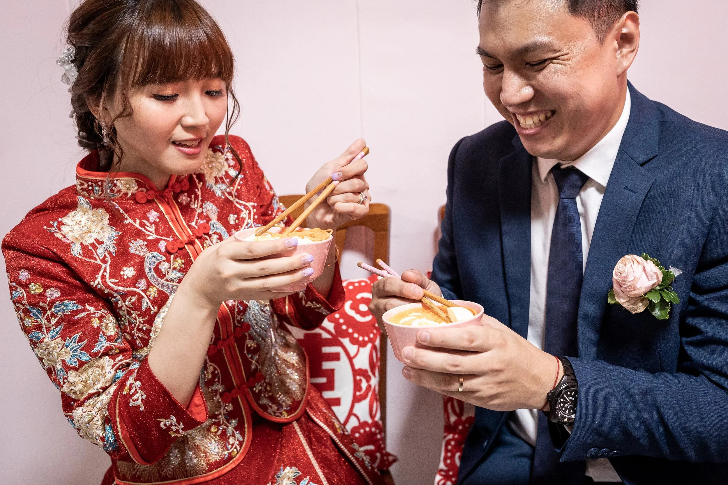 A bride in a traditional red Chinese wedding dress shares a bowl of noodles with a groom in a blue suit, both smiling.