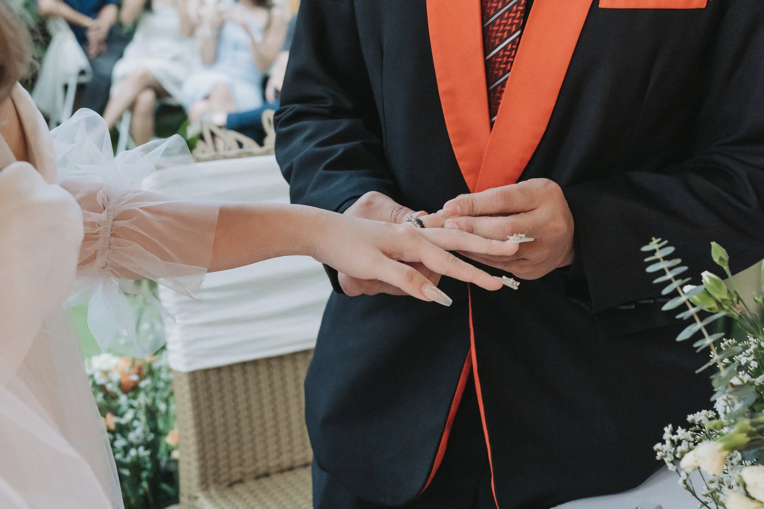 Close-up of a wedding ceremony where a bride places her hand on the groom's hand, both are dressed formally, with the groom in a dark suit with orange accents.