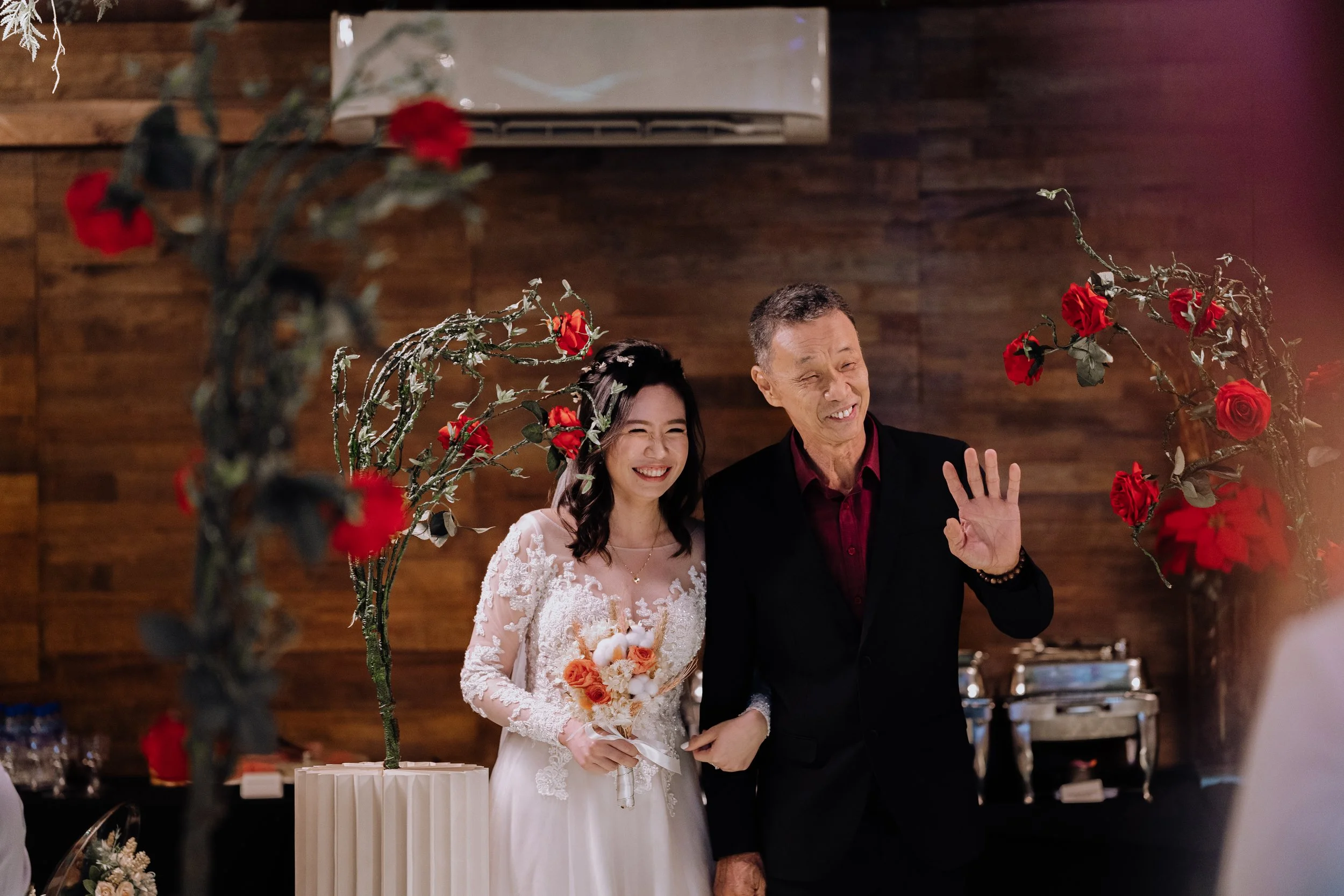 A bride and an older man, possibly her father, are smiling and walking down the aisle at a wedding. The bride wears a white lace wedding dress and holds a bouquet, while the man wears a black jacket and a red shirt. They are framed by decorative bran