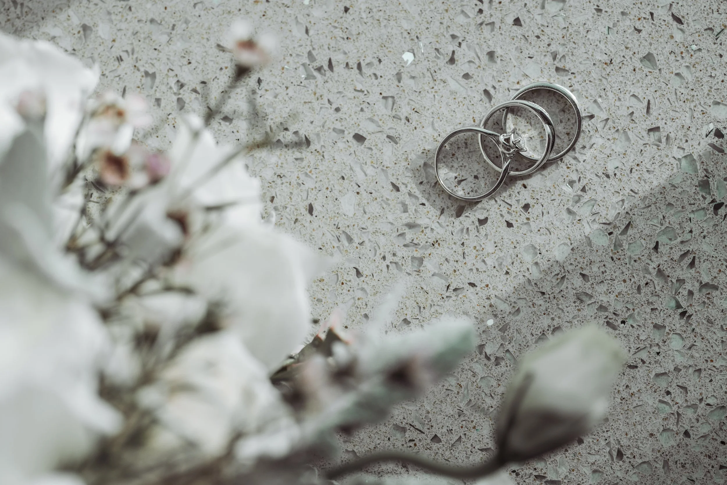 A pair of silver wedding rings placed on a speckled beige countertop with white flowers in the foreground.