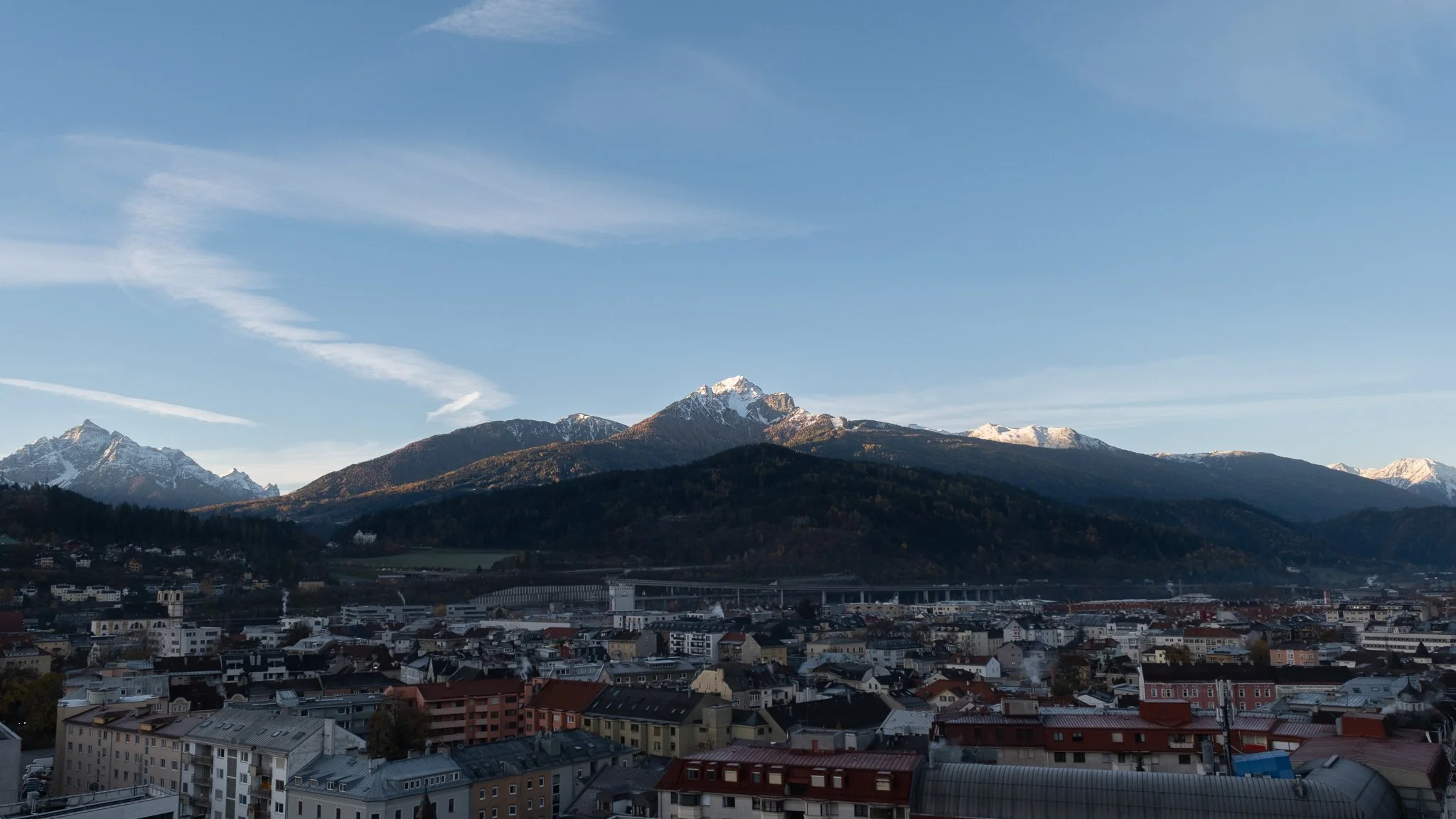 Cityscape with buildings in the foreground, mountainous landscape in the background, snow-capped peaks, and a clear blue sky.