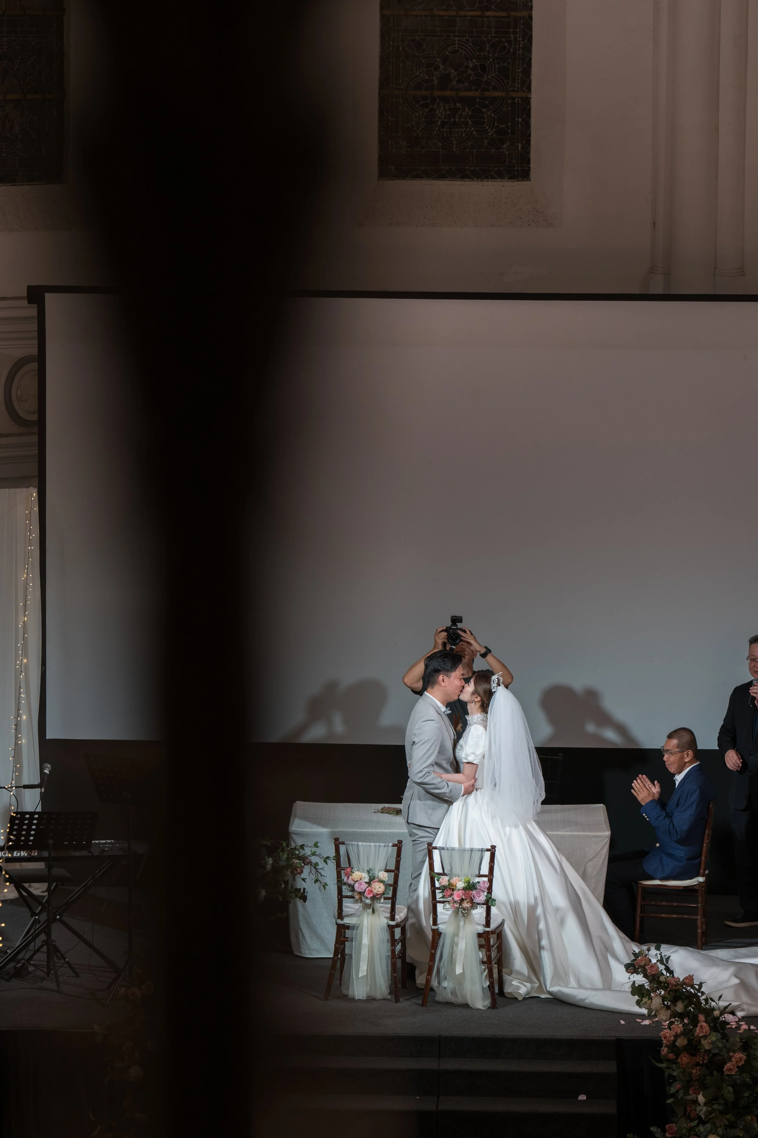 A wedding ceremony with a bride and groom kissing on stage, surrounded by family and friends, with a person taking photos in the background.