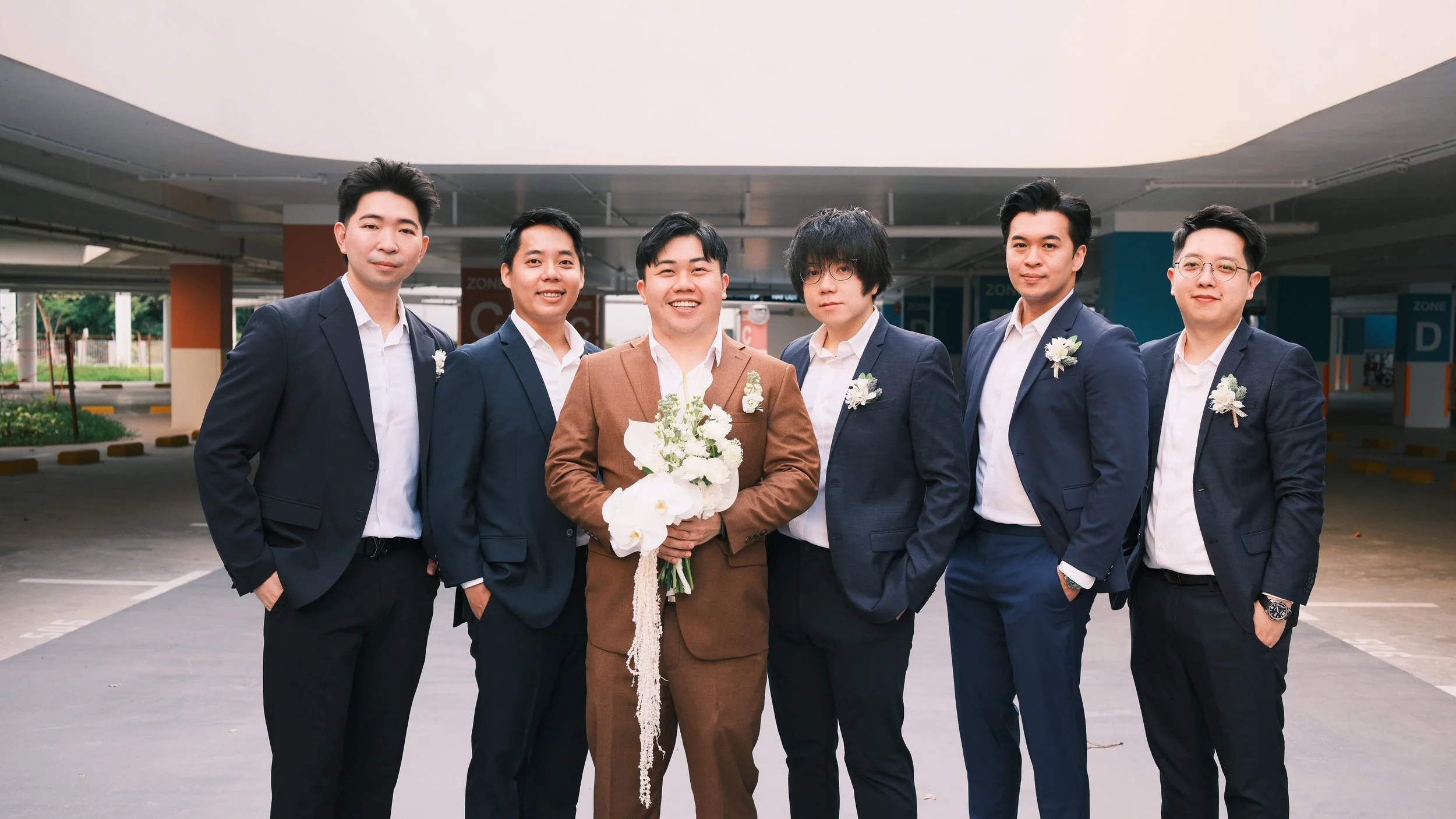 Group of six men dressed in suits standing in an outdoor parking lot, with the man in the center holding a bouquet of white flowers, likely at a wedding or formal event.