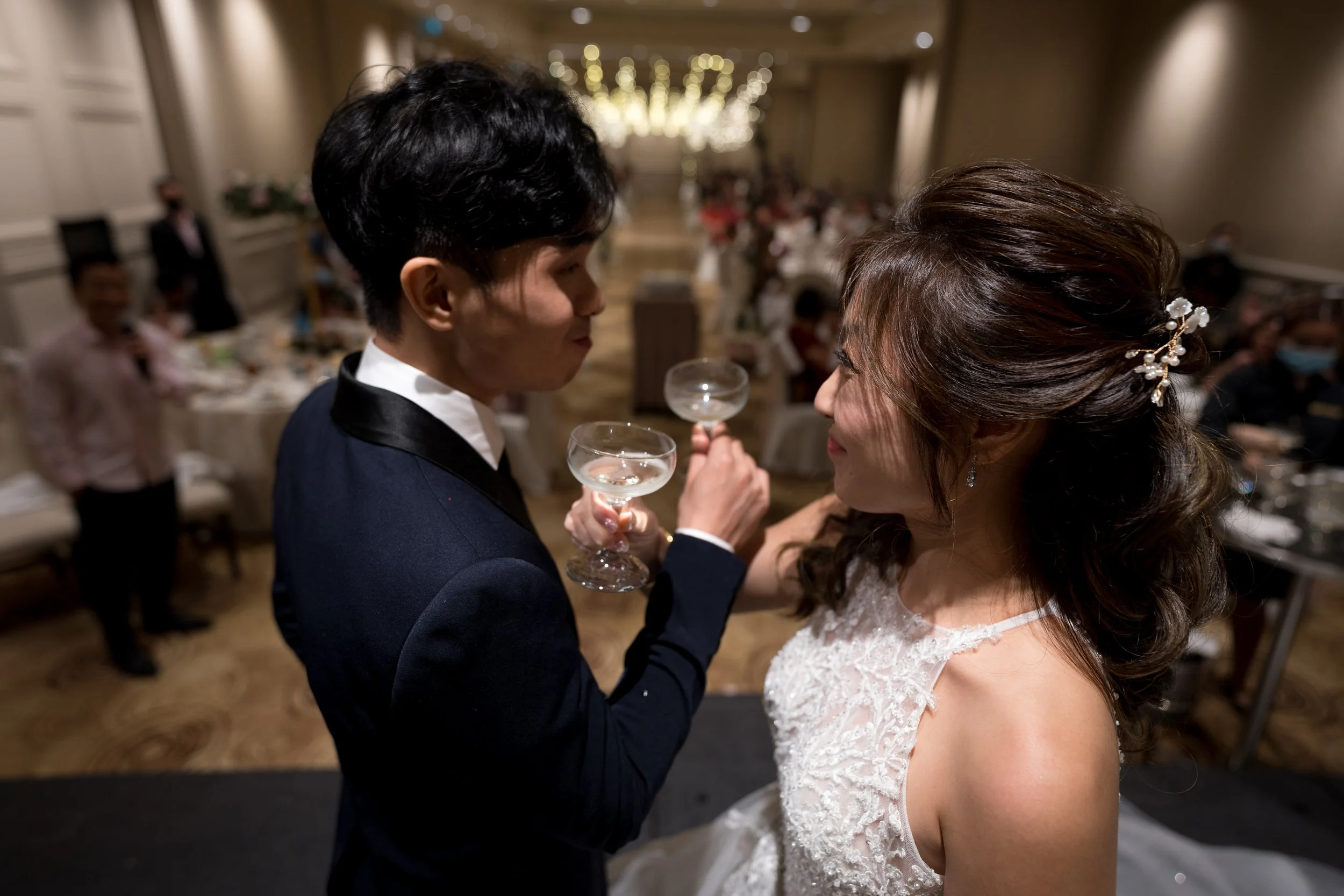 A couple sharing a toast at a wedding reception, with the groom in a tuxedo and the bride in a wedding dress, facing each other and smiling.