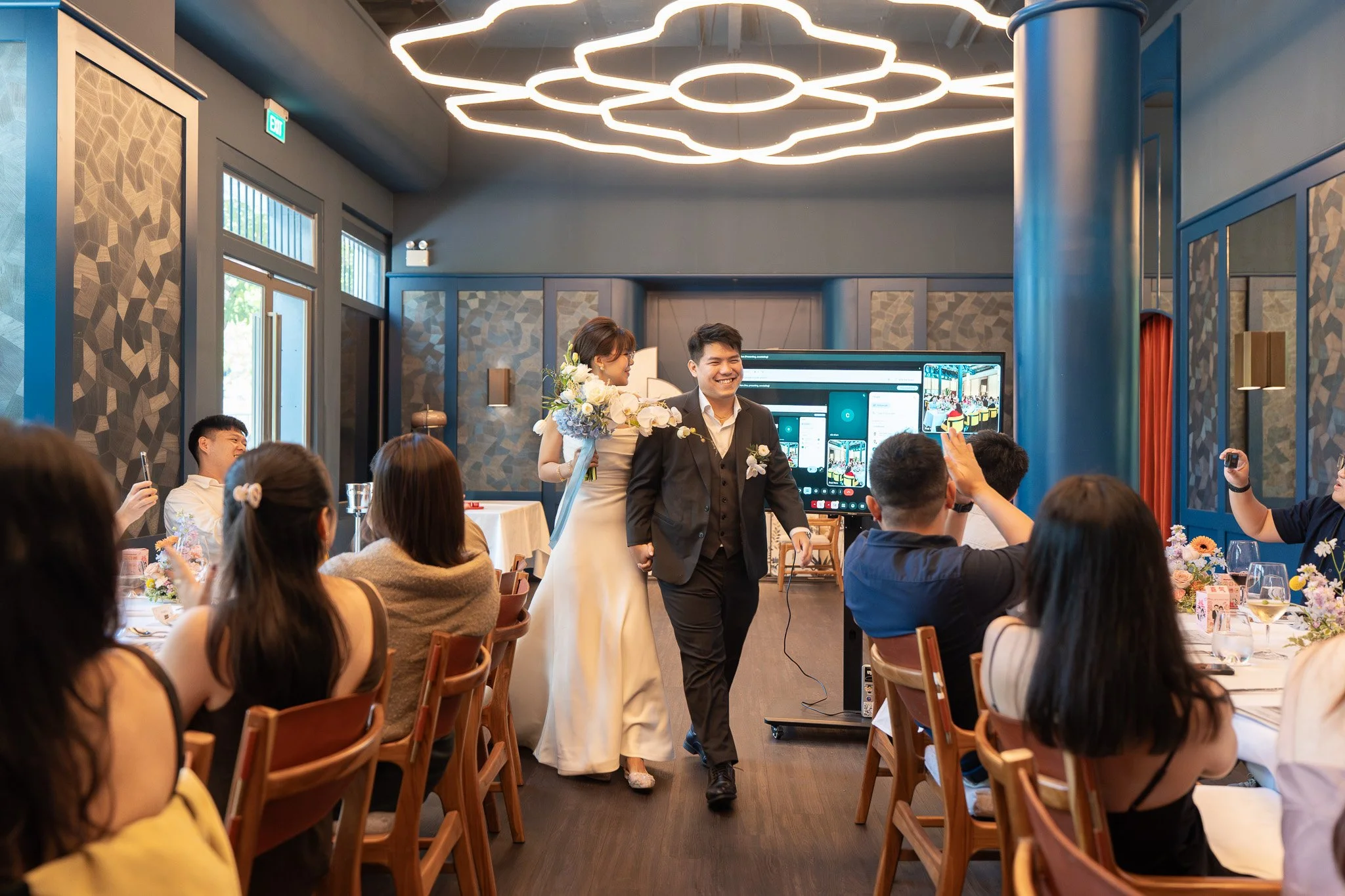 A wedding reception with a bride and groom entering a decorated banquet room, surrounded by seated guests taking photos.
