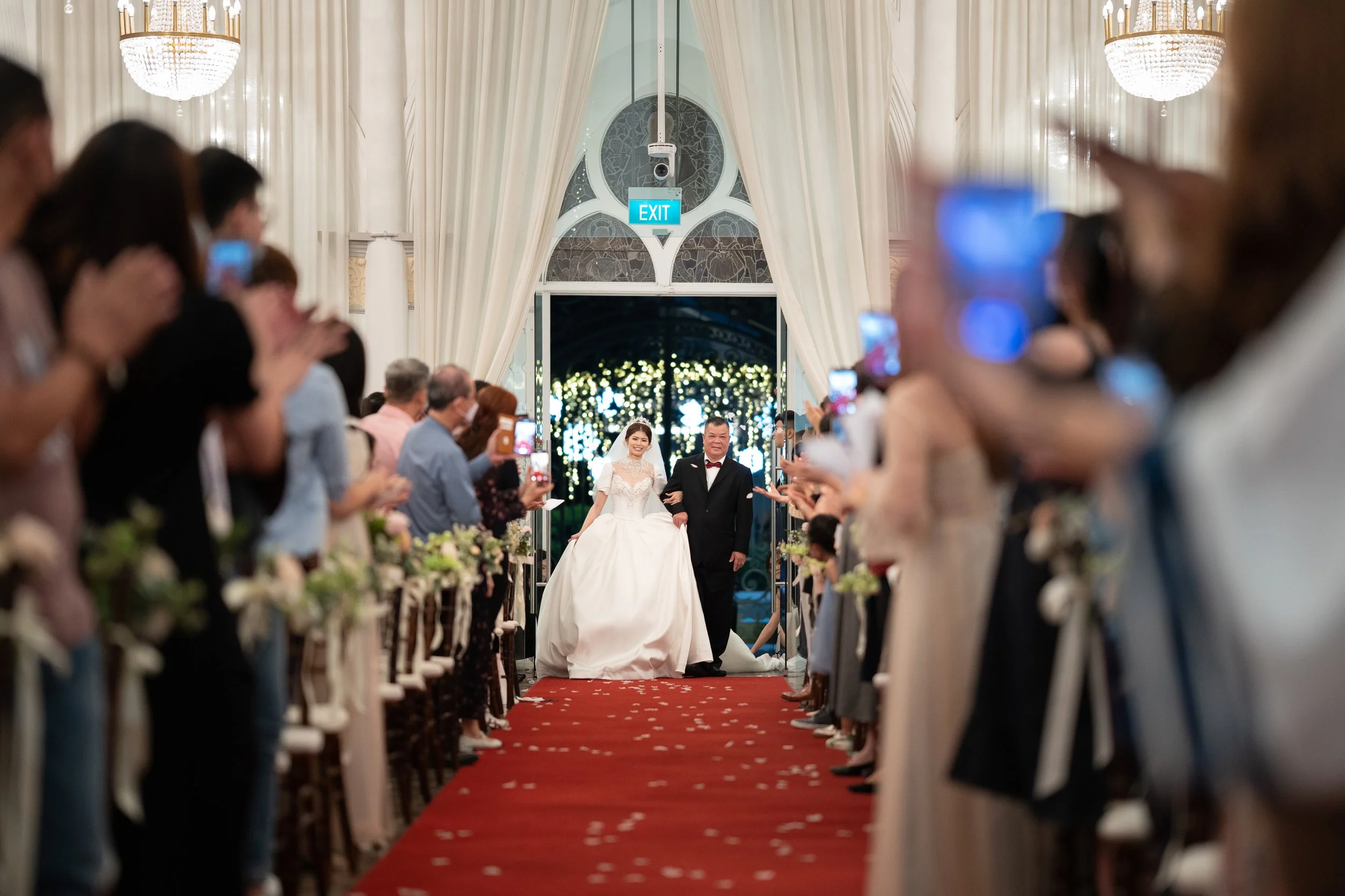 A bride in a white wedding gown and a man in a tuxedo walking down the aisle of a wedding ceremony, surrounded by seated guests taking photos, with a decorated backdrop behind them and white curtains framing the aisle.