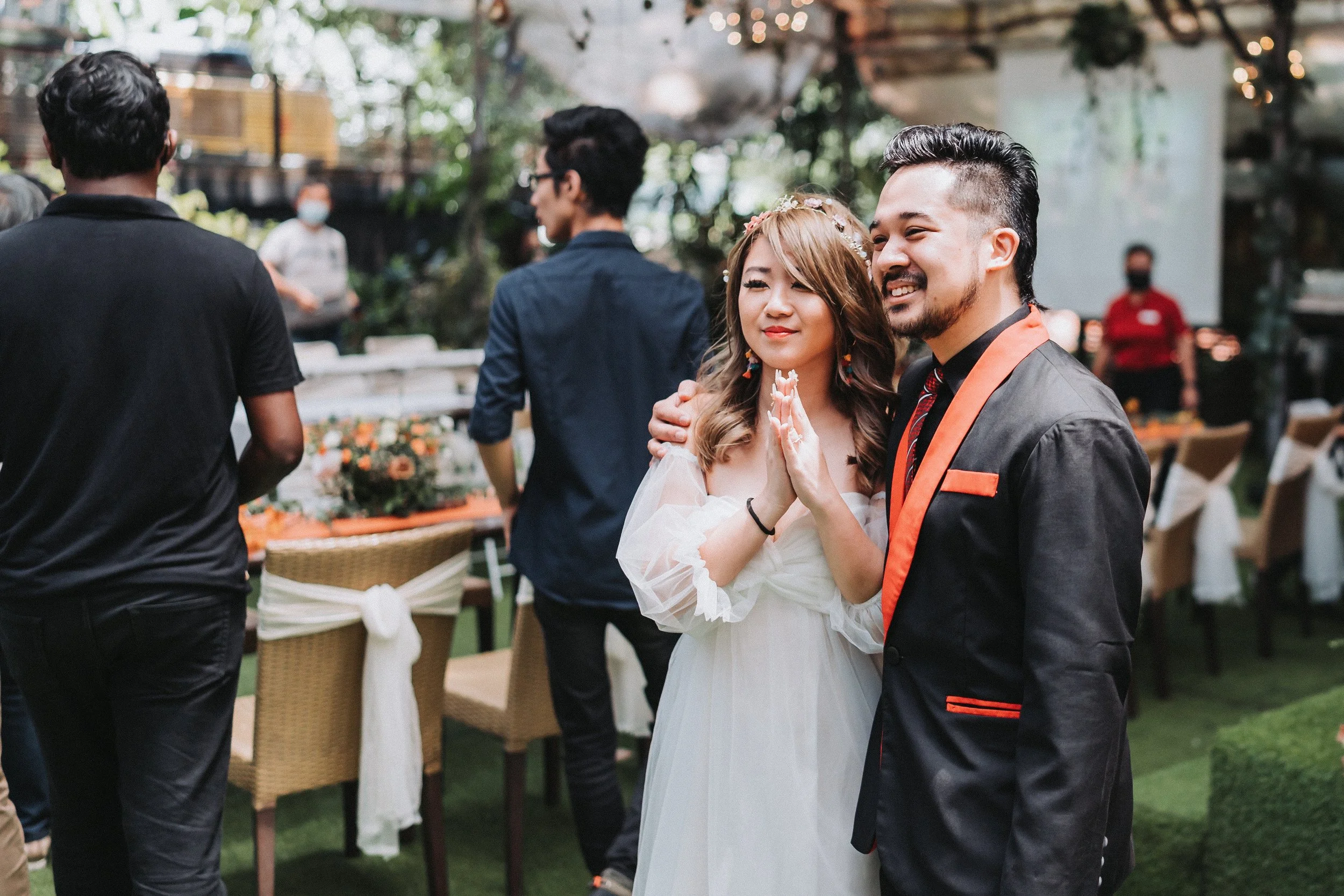 A happy couple, dressed in formal wedding attire, stands outdoors at a garden wedding reception. The woman is in a white dress with a floral crown, and the man is in a black suit with orange accents. They are smiling and holding hands, with wedding g