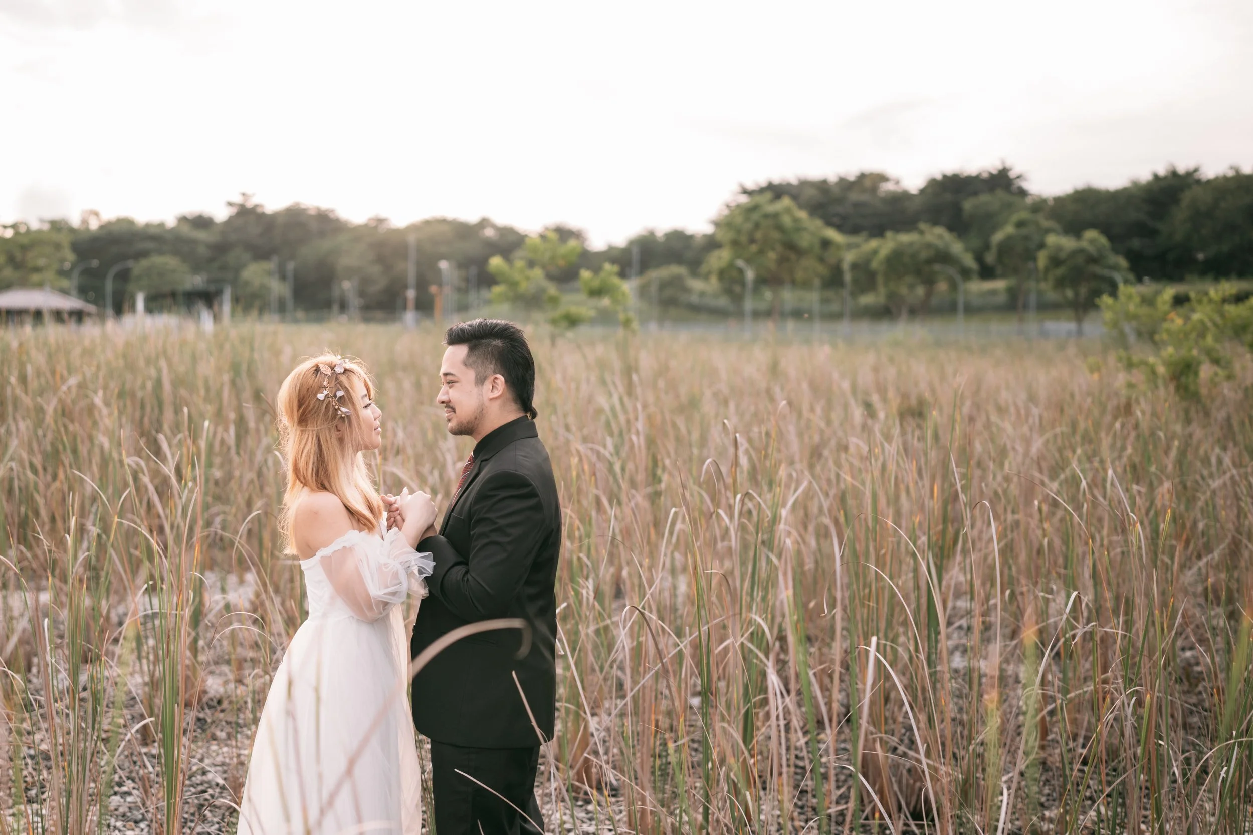A couple holding hands and looking at each other in a grassy field during sunset, with trees in the background.